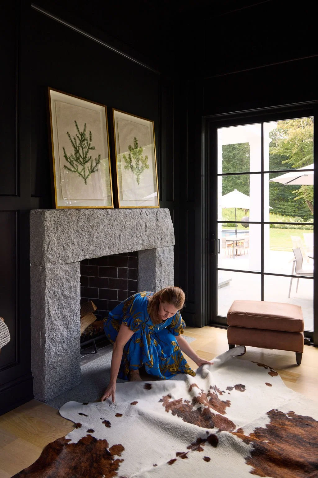 A woman in a blue dress is kneeling on the floor, arranging a cowhide rug in front of a gray stone fireplace in a living room with black walls. Two botanical illustrations are hanging above the fireplace, and a sliding glass door leads outside to a patio with umbrellas and outdoor furniture.