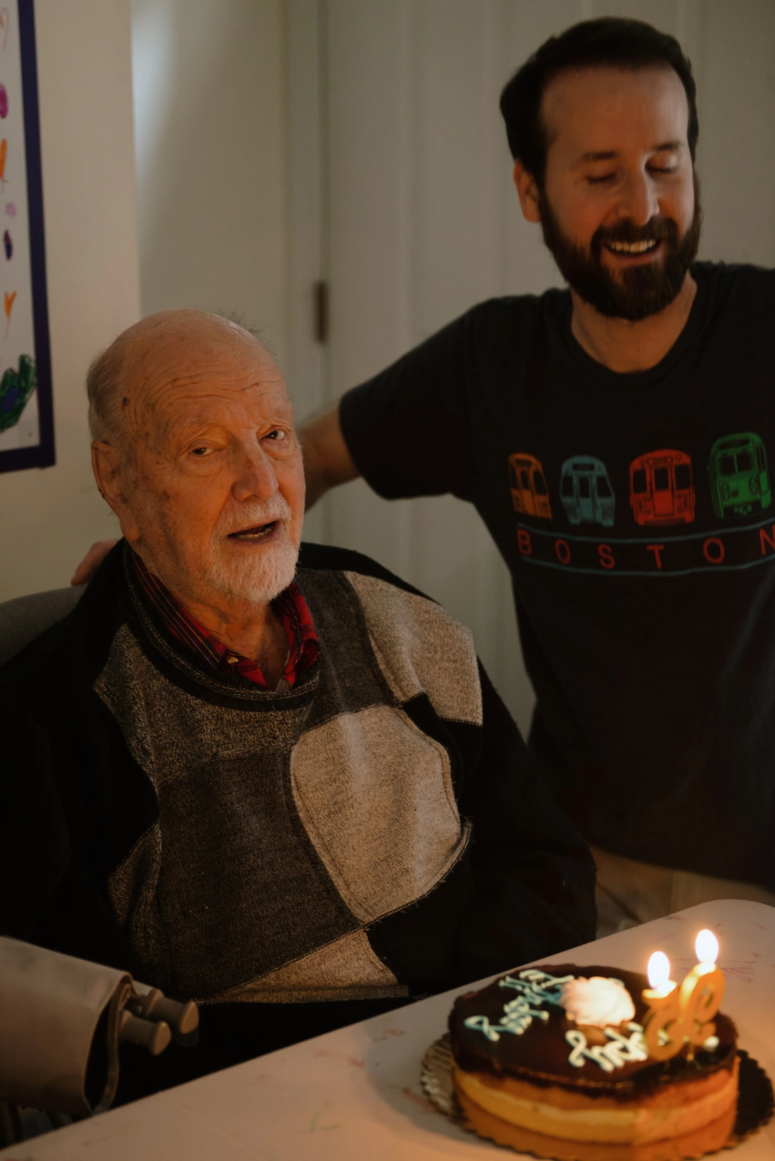 An elderly man with gray hair and a beard celebrating his birthday with a cake, candles, and a younger man standing next to him smiling.
