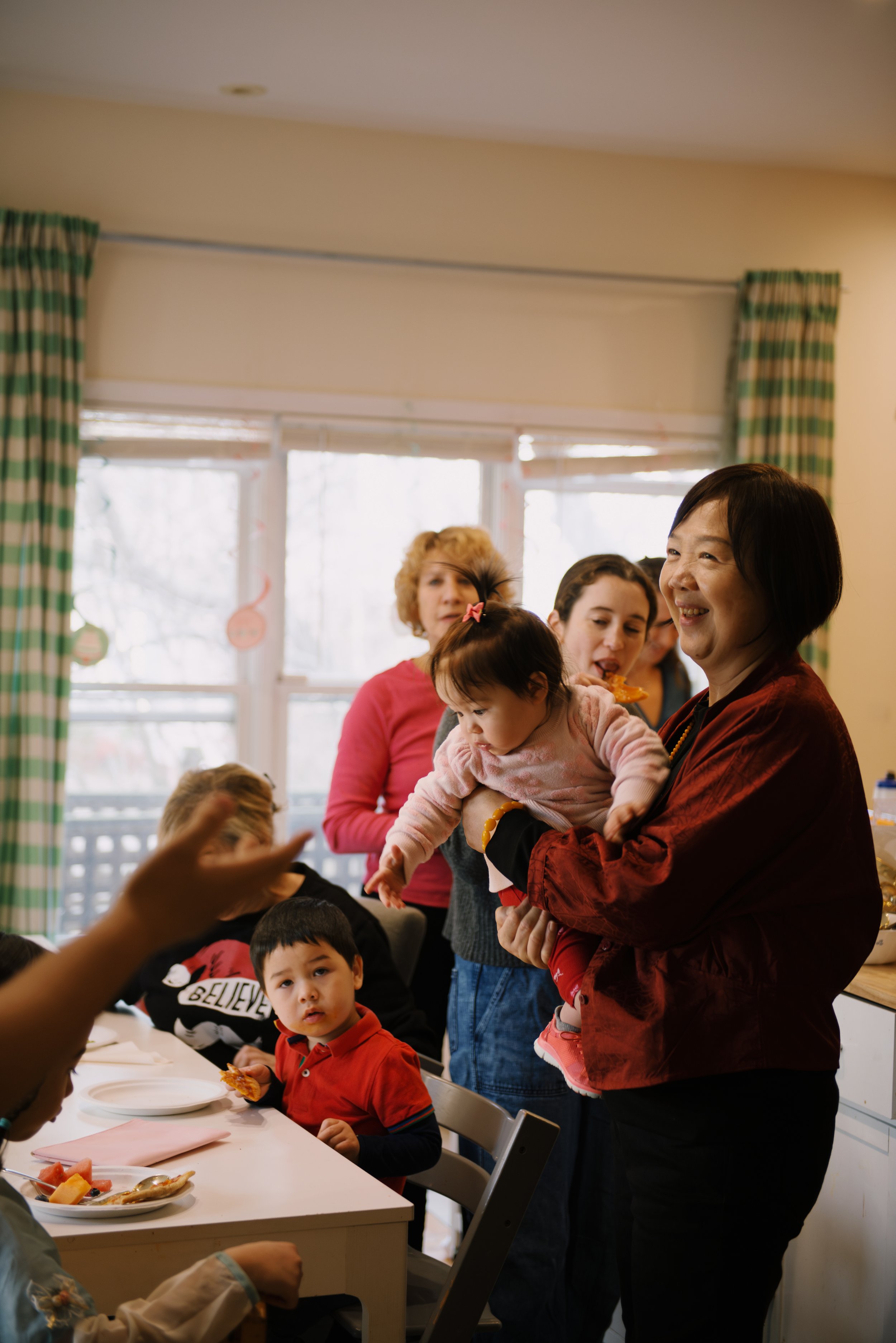 Family gathering around a dining table during a celebration in a cozy home, with a woman holding a young child and other family members interacting.