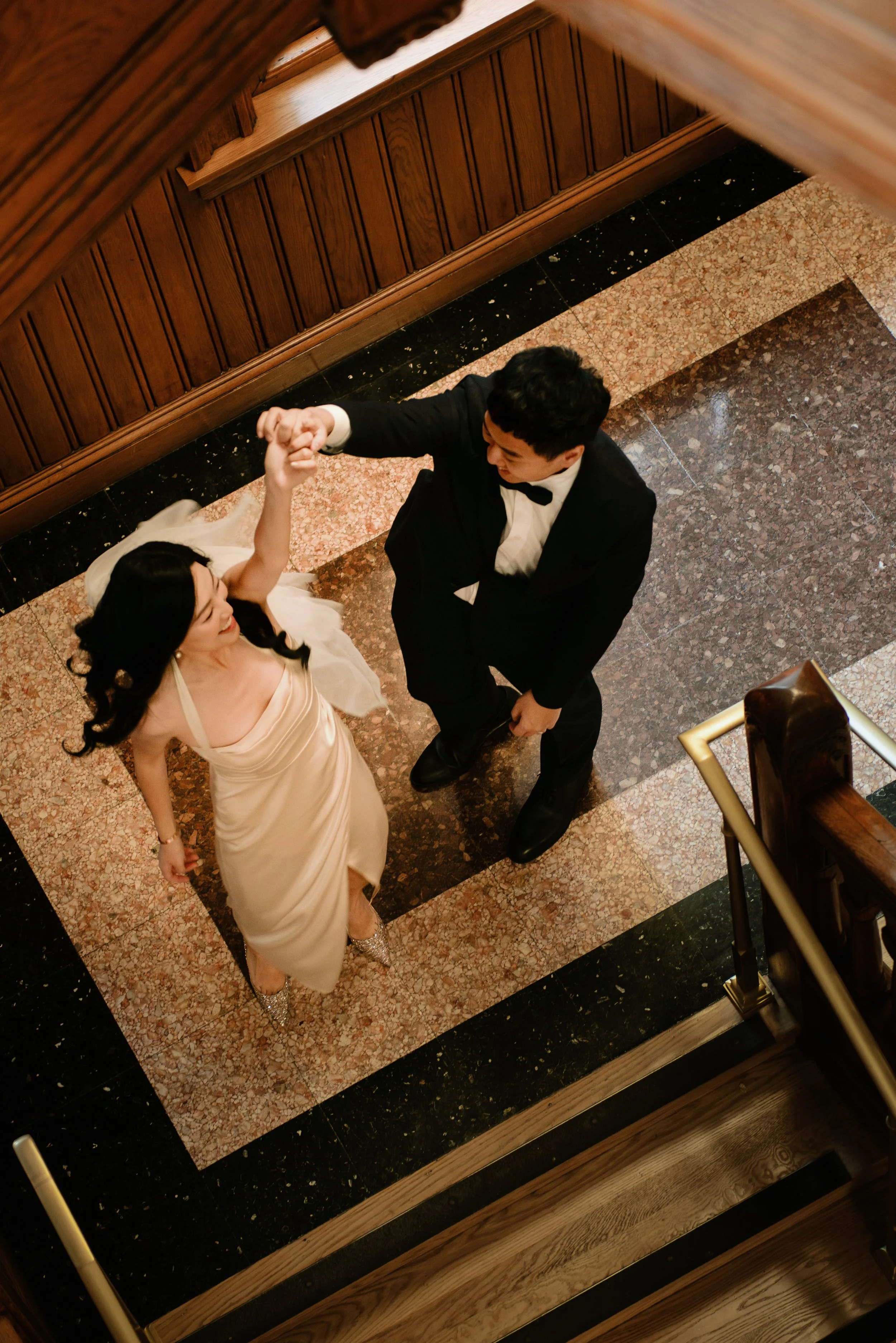 A couple dancing on a patterned floor, viewed from above. The woman wears a cream-colored dress and sparkling heels, while the man is in a black tuxedo with a bow tie.