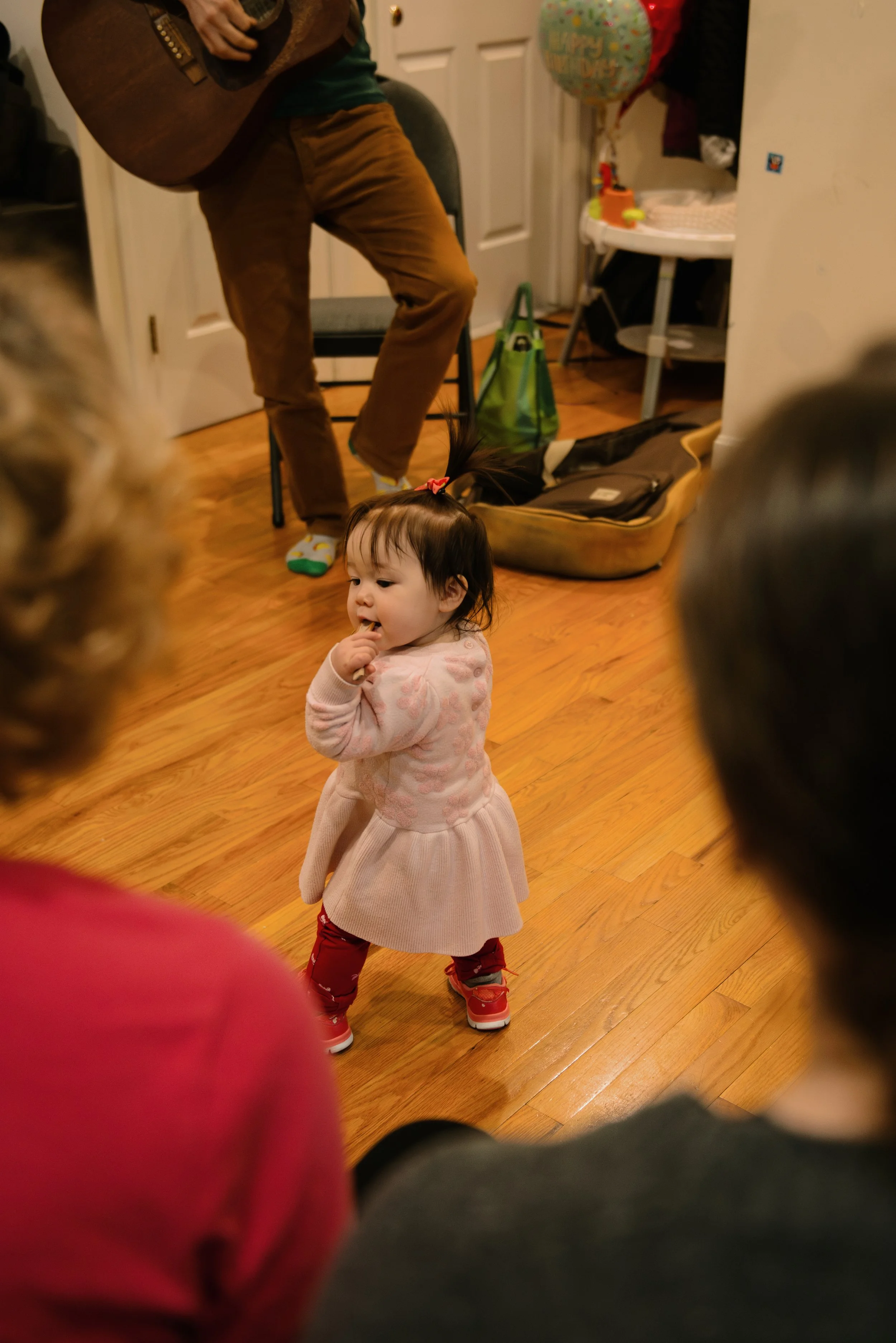 A young girl in a pink dress, red pants, and red shoes is standing on a hardwood floor, looking to her right with her finger in her mouth. Several adults, partially visible, are standing nearby, and a man is playing a guitar in the background. The ro