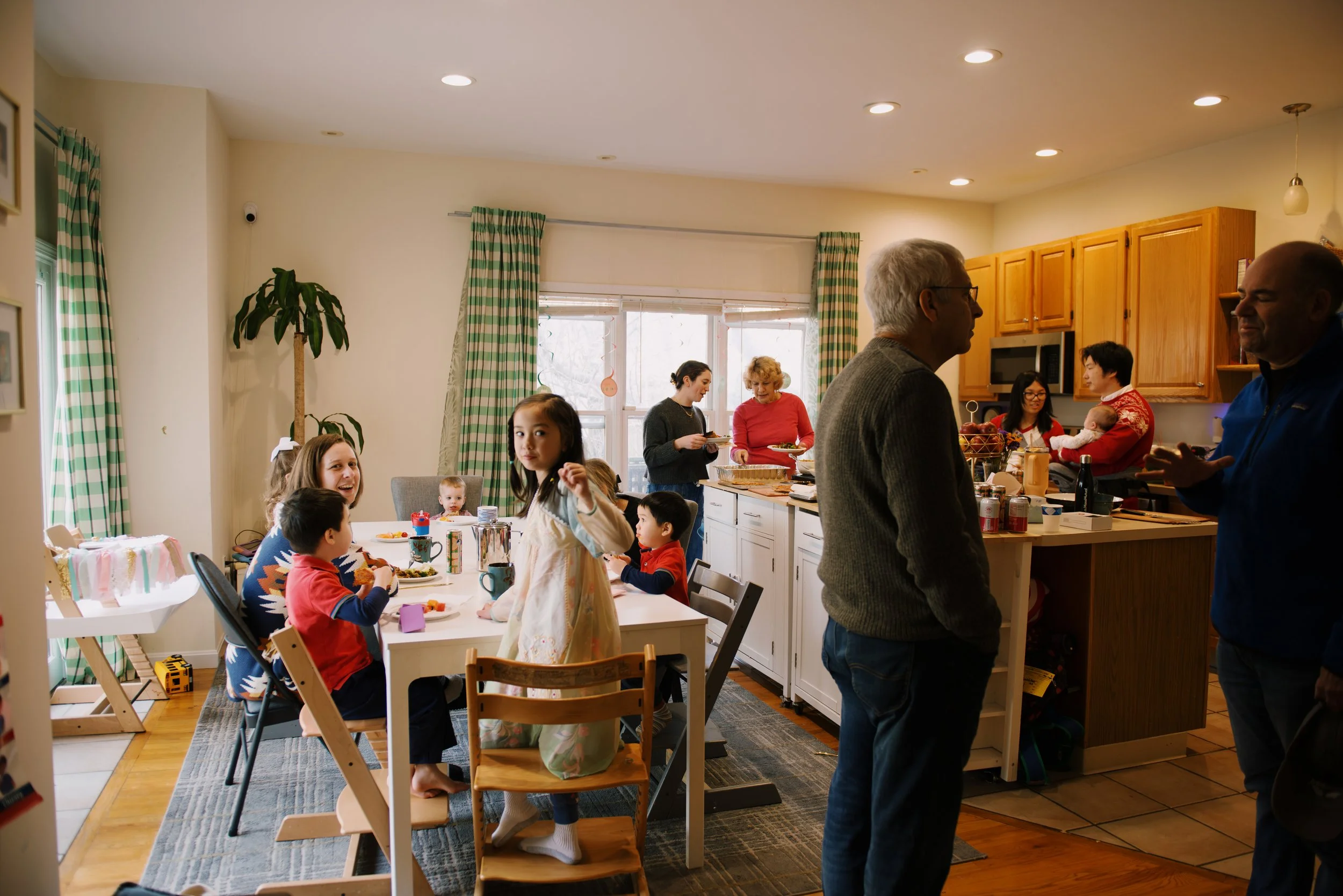 Family gathering in kitchen during daytime, with children at the dining table and adults engaged in conversation.