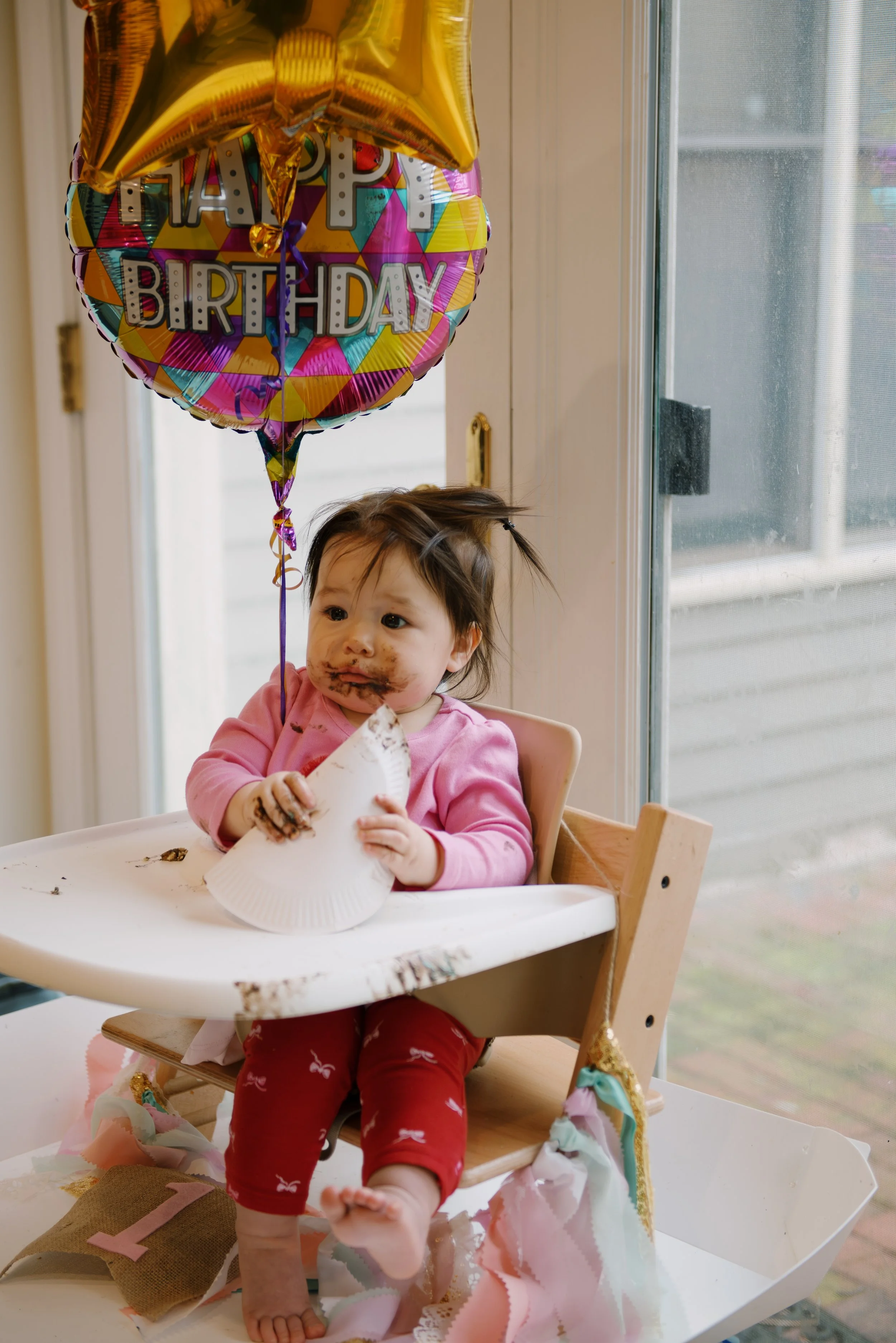 Baby girl with messy chocolate face and hands, sitting in a high chair holding a paper plate, with a 'Happy Birthday' balloon floating above her at a birthday celebration.