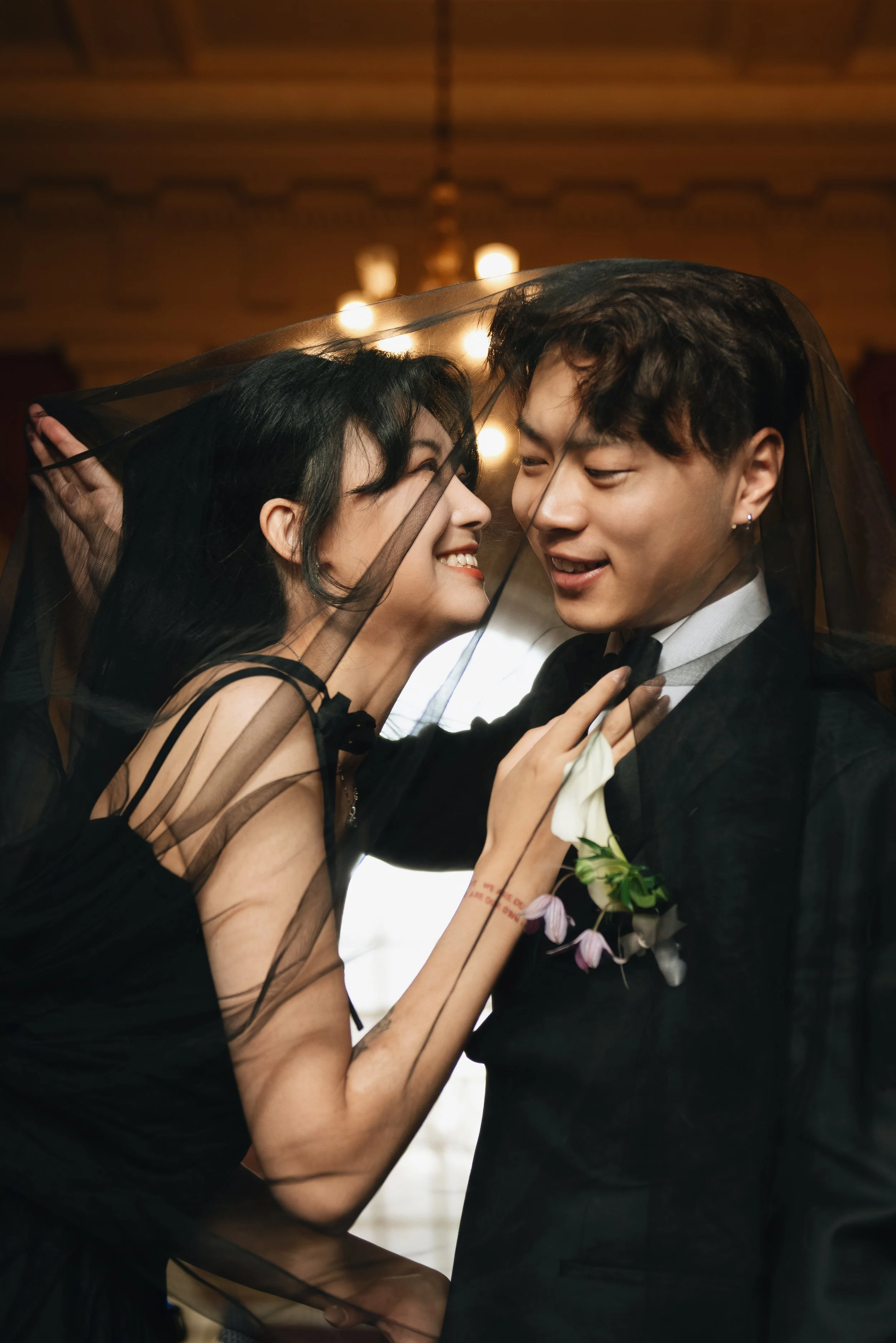 A couple shares an intimate moment under a veil, smiling at each other in an elegant setting, possibly during a wedding or formal event.
