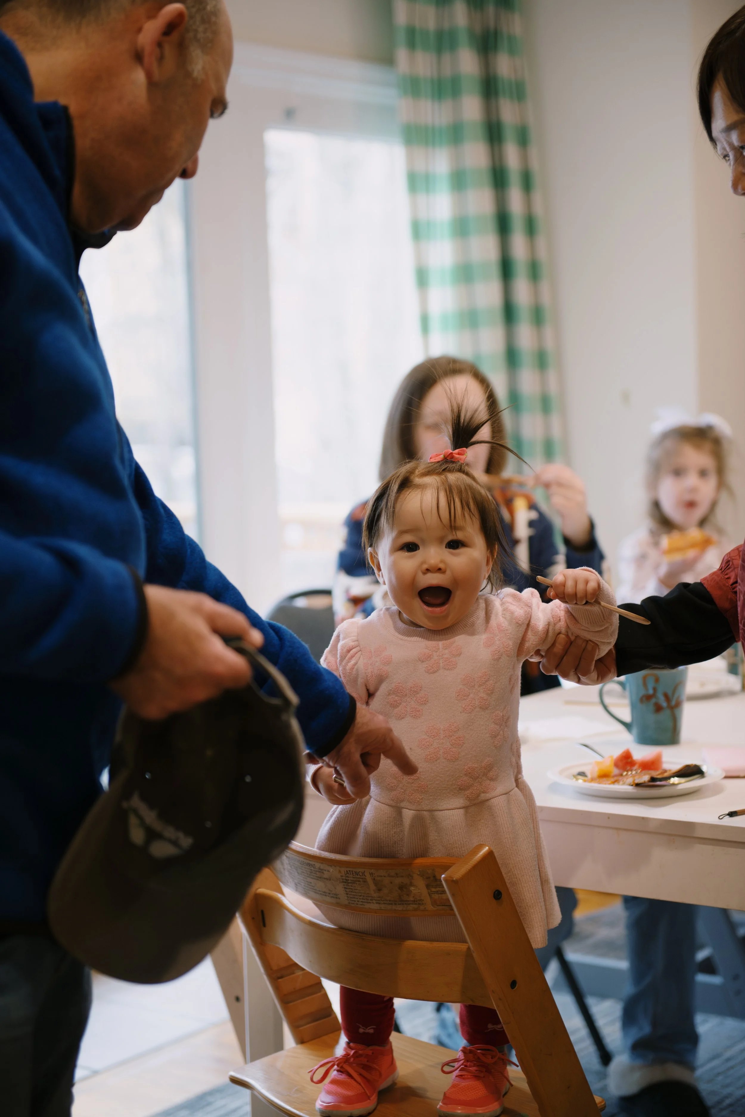 A young girl with a surprised expression standing on a high chair at a table during a meal, with adults nearby. The scene appears to be in a bright, homey dining area.