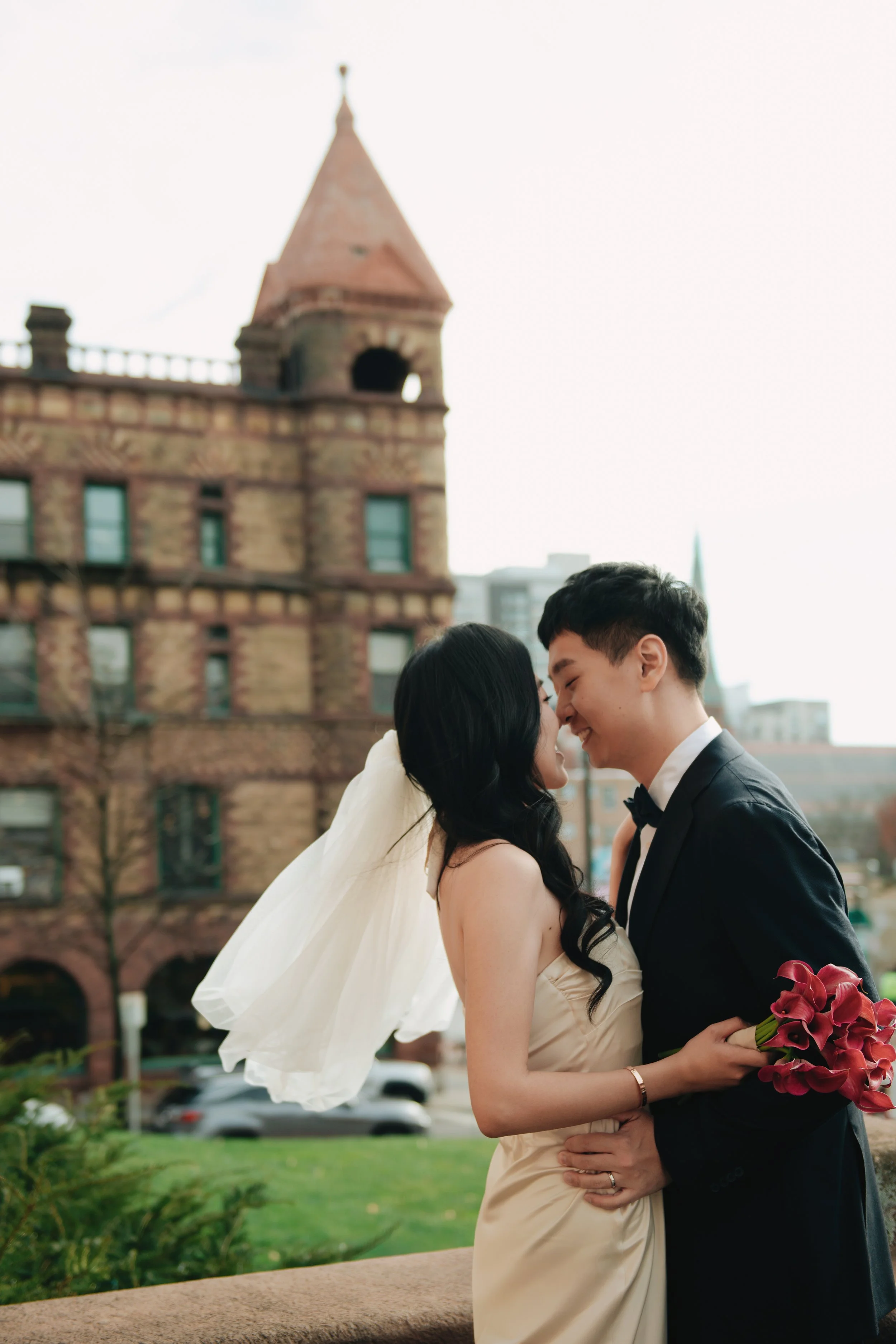 A bride and groom are smiling and touching noses, standing outdoors in front of a historic building with a tower.