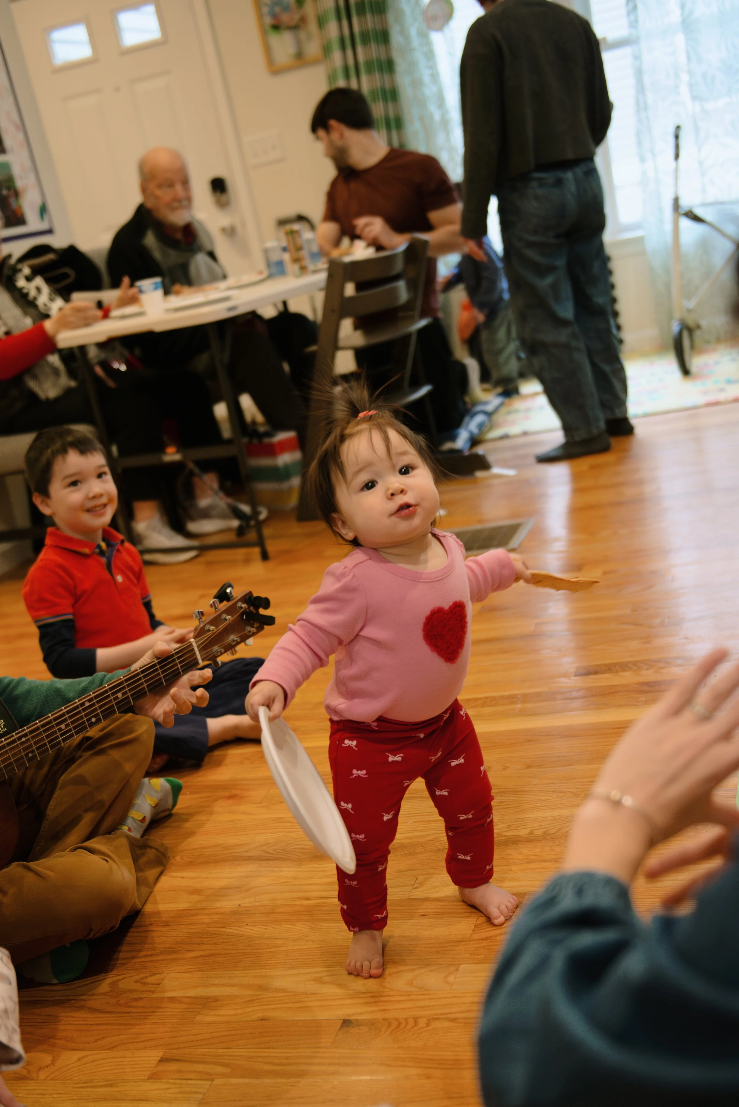 A young girl with a pink shirt with a red heart on it, red leggings with white birds, standing on a wooden floor, holding a paper plate and a paper. Behind her, a boy in a red and navy shirt sitting on the floor and smiling, and a man playing a guita