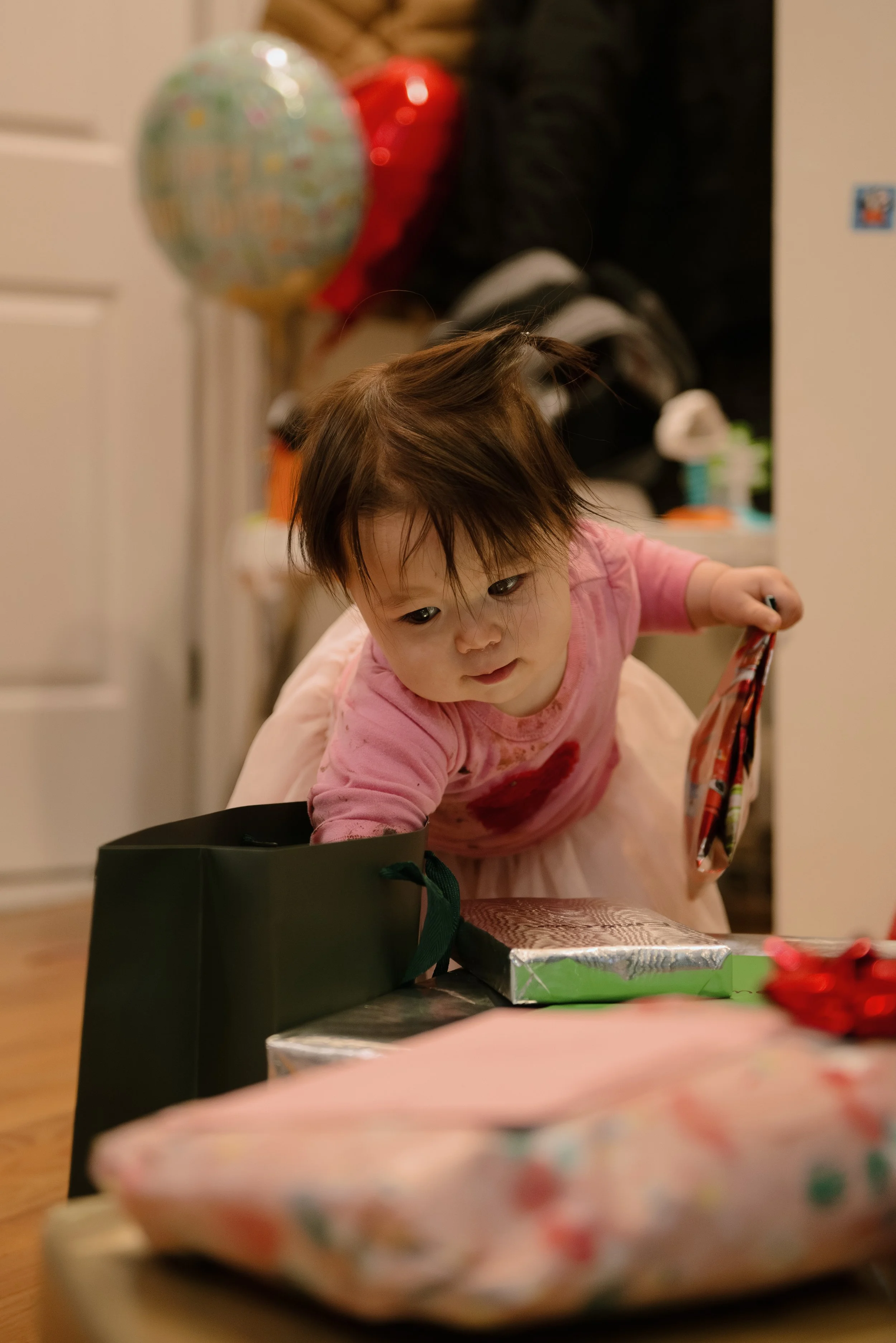 A young girl with dark hair and a pink shirt leaning over a table with wrapped presents, holding a small item in her hand, with balloons in the background.