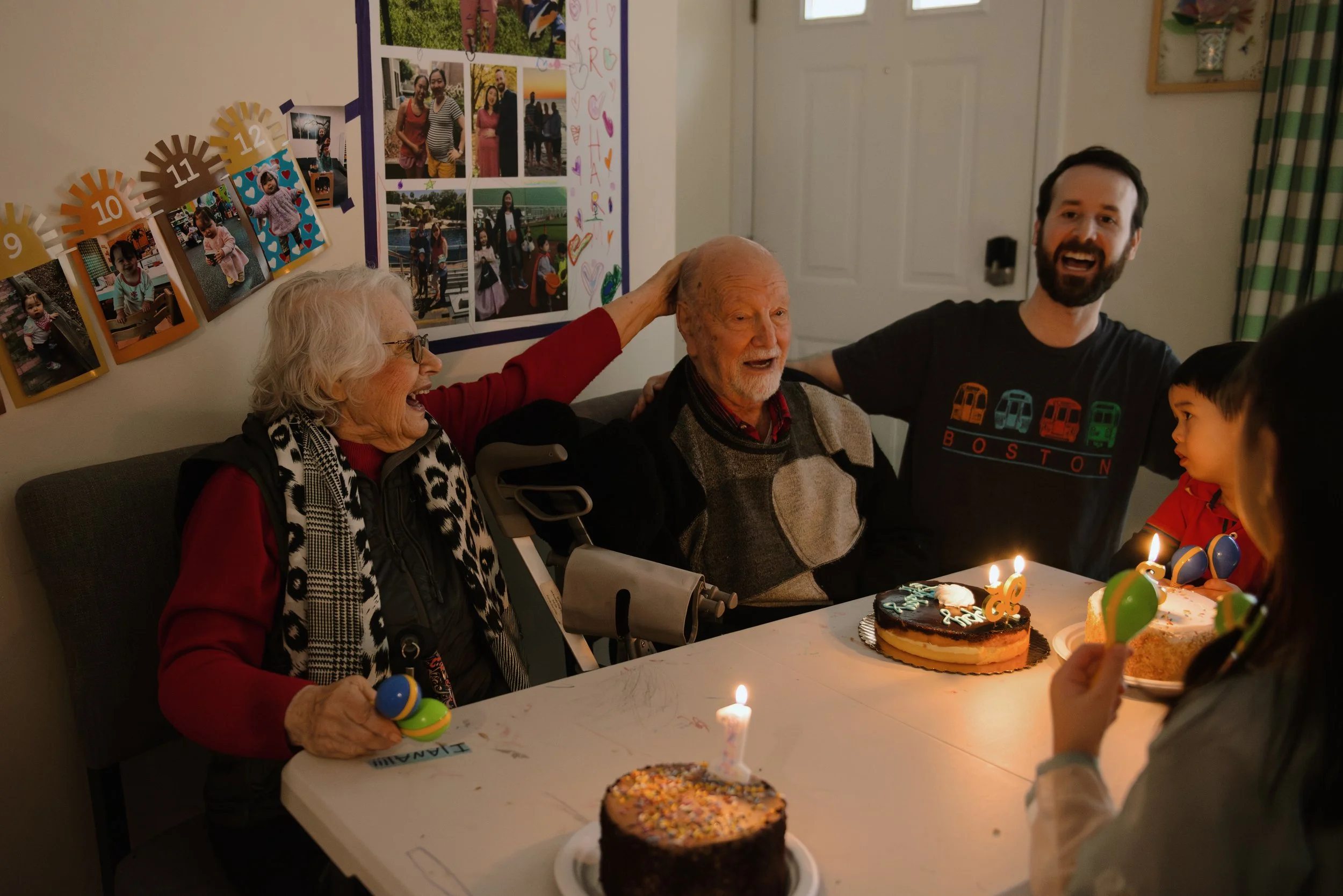 People celebrating a birthday, with an elderly man sitting at the center, surrounded by family members who are smiling and laughing. There are two birthday cakes with lit candles on the table, and children holding balloons. The room has birthday deco