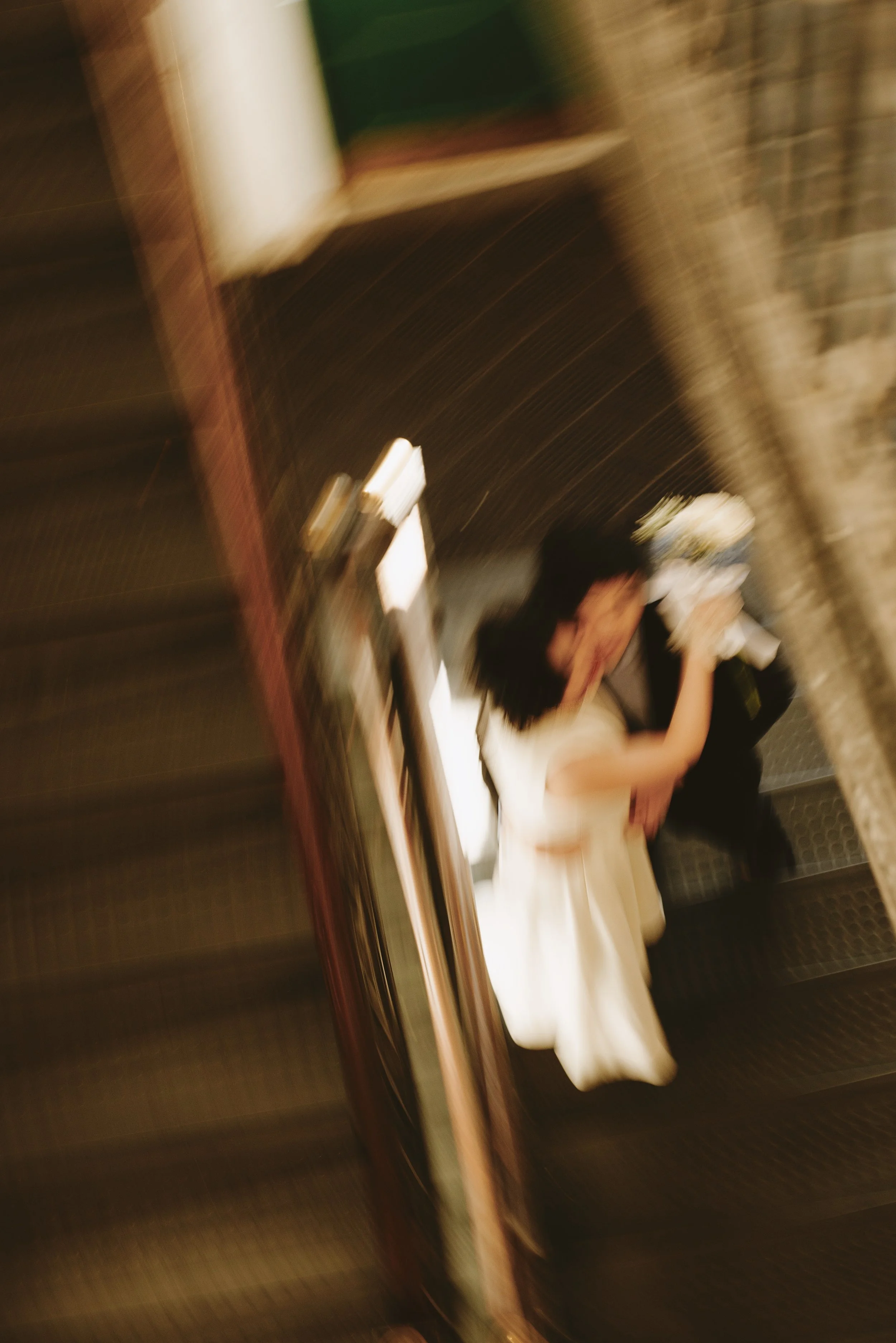 A blurry photo of a couple on a staircase, with the woman in a white dress and the man in dark clothing, embracing and holding a bouquet.