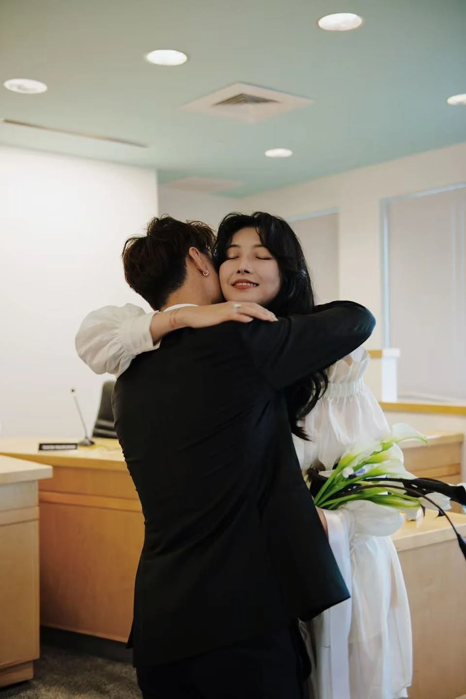 A woman in a white dress hugging a man in a black suit inside an office or reception area, holding white calla lilies, both smiling with closed eyes.