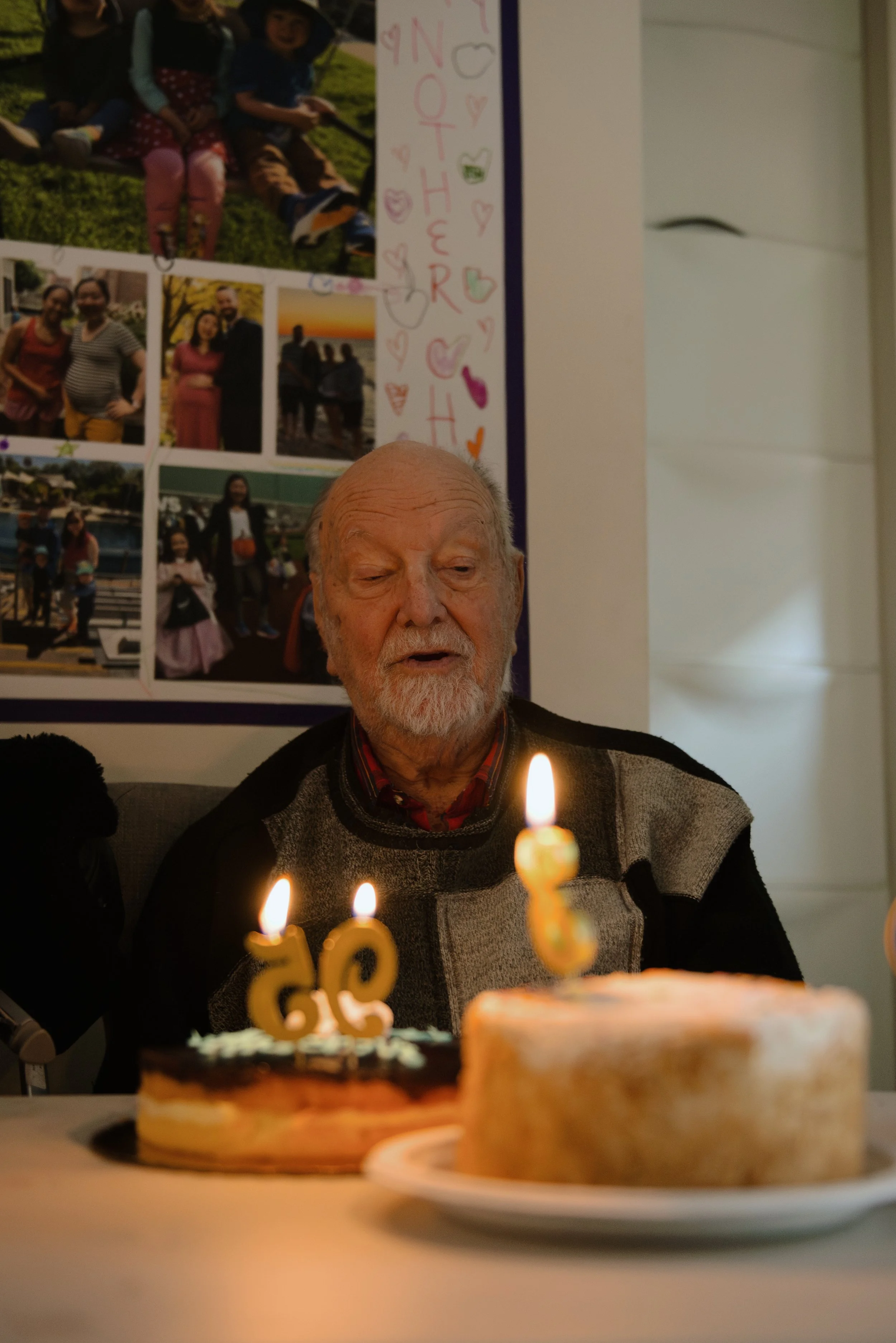 An elderly man with a white beard sitting at a table with two birthday cakes, one with a candle shaped as '86', celebrating his 86th birthday.