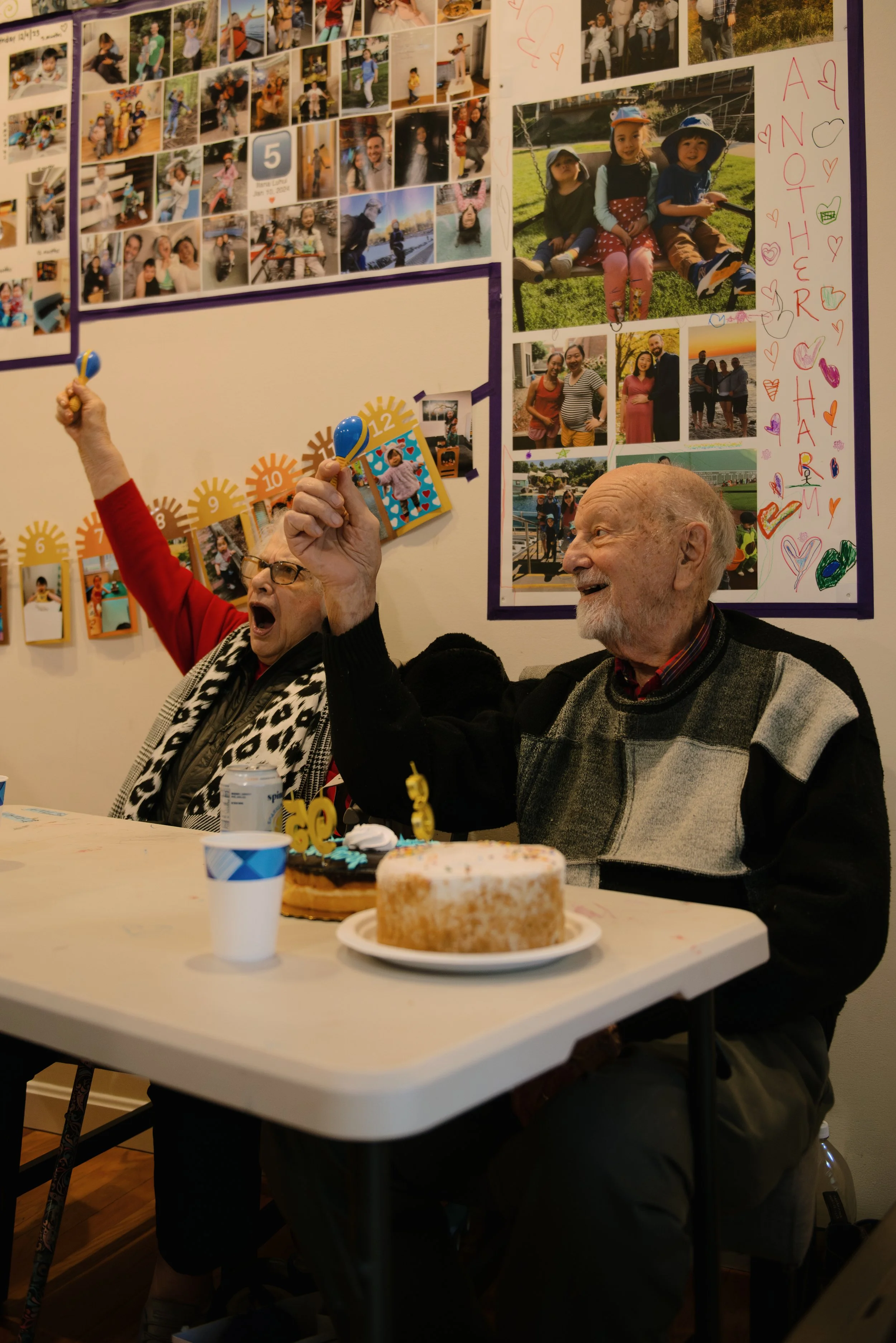An elderly man and woman celebrating at a birthday party; the woman is holding a toy maraca and the man is smiling, with birthday cakes and party decorations on the table. The background features a wall with a collage of photos and colorful crafts.