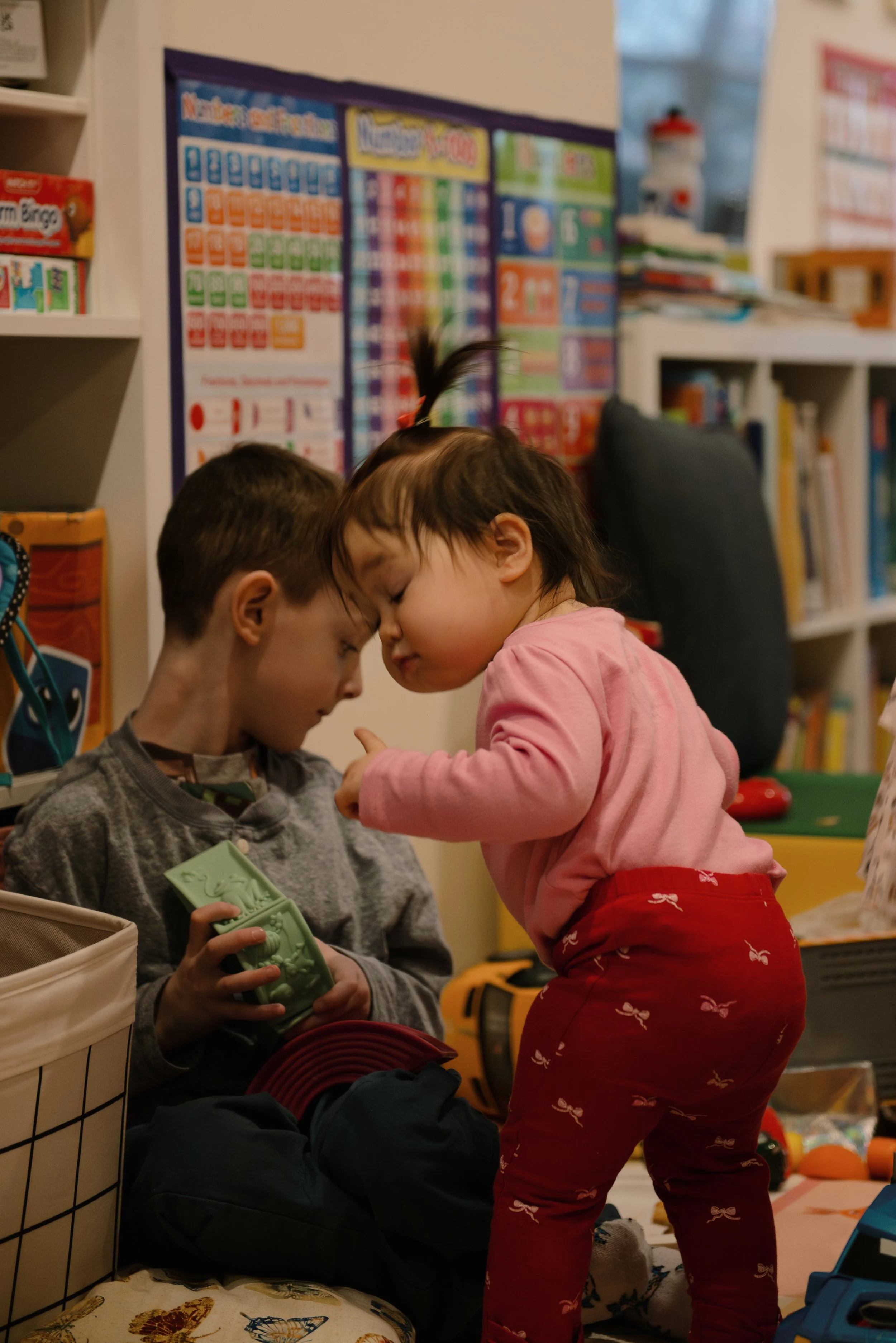 Two young children, a boy and a girl, are sharing a close moment with their foreheads touching inside a classroom filled with educational posters and books.