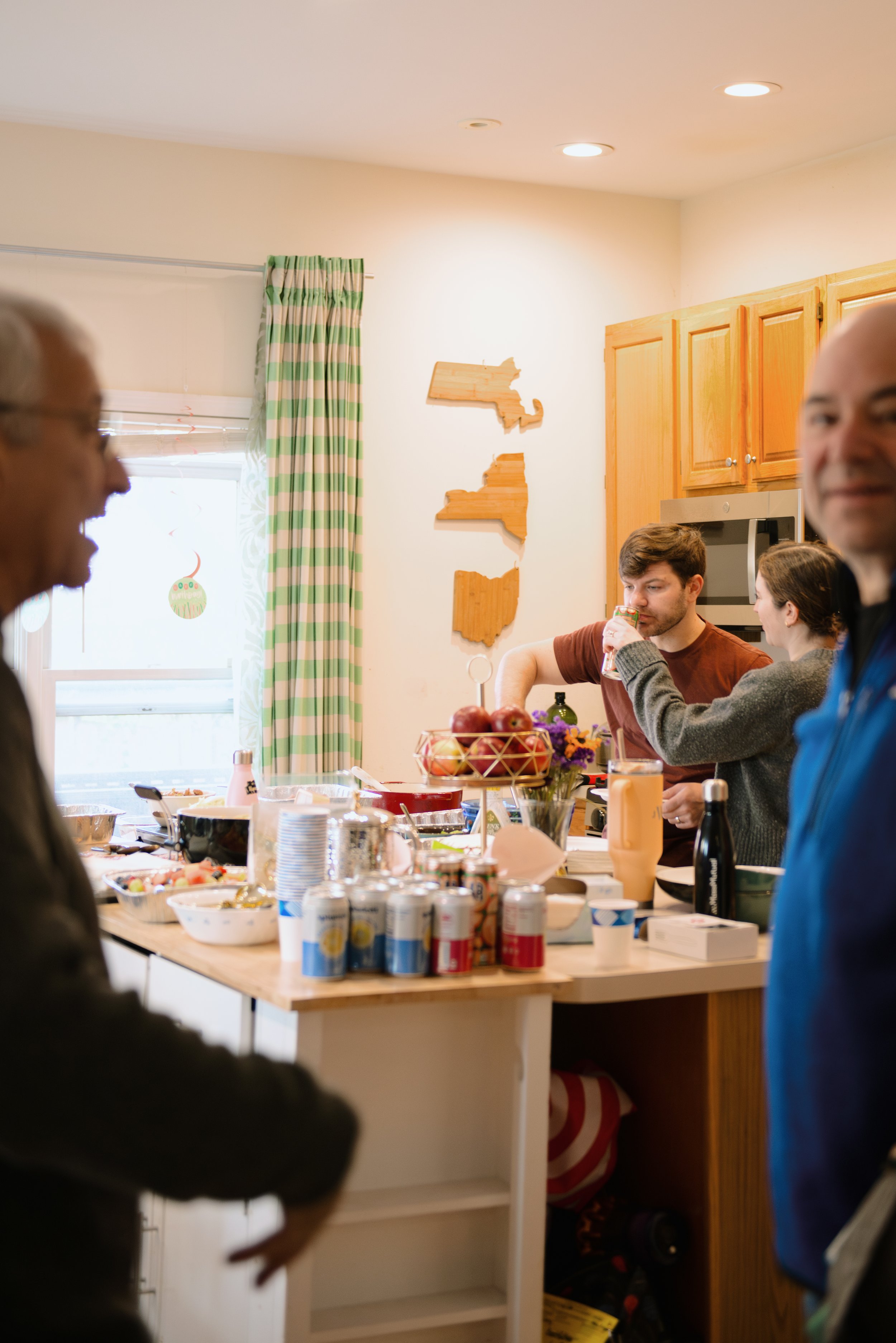 Family gathering in a kitchen with people around a cluttered breakfast table, Christmas decorations visible outside the window, wooden wall art of the state of Massachusetts.