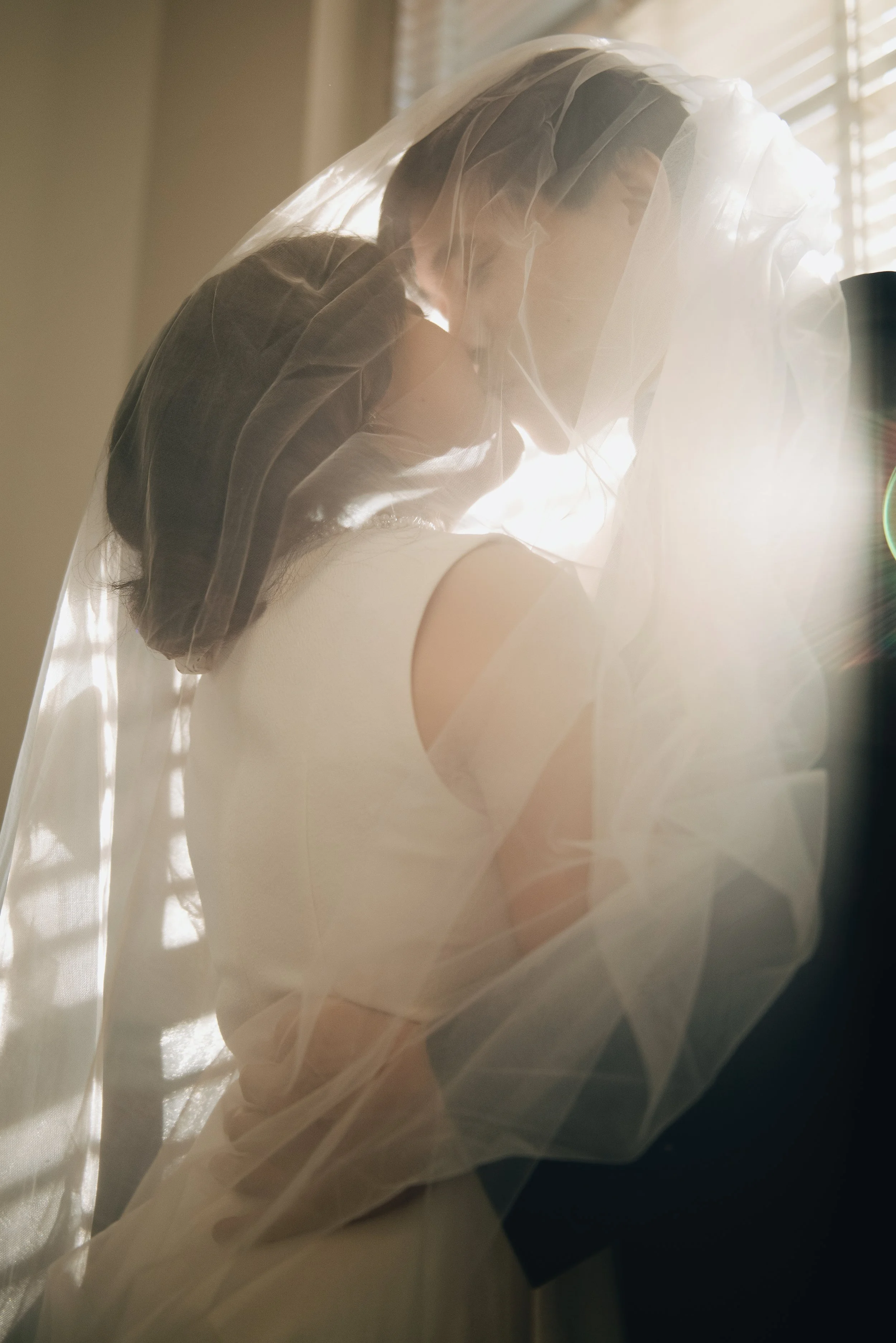 A bride with dark hair and light skin, dressed in a white wedding gown, sitting by a window with sunlight shining through blinds, wearing a veil draped over her head and shoulders.