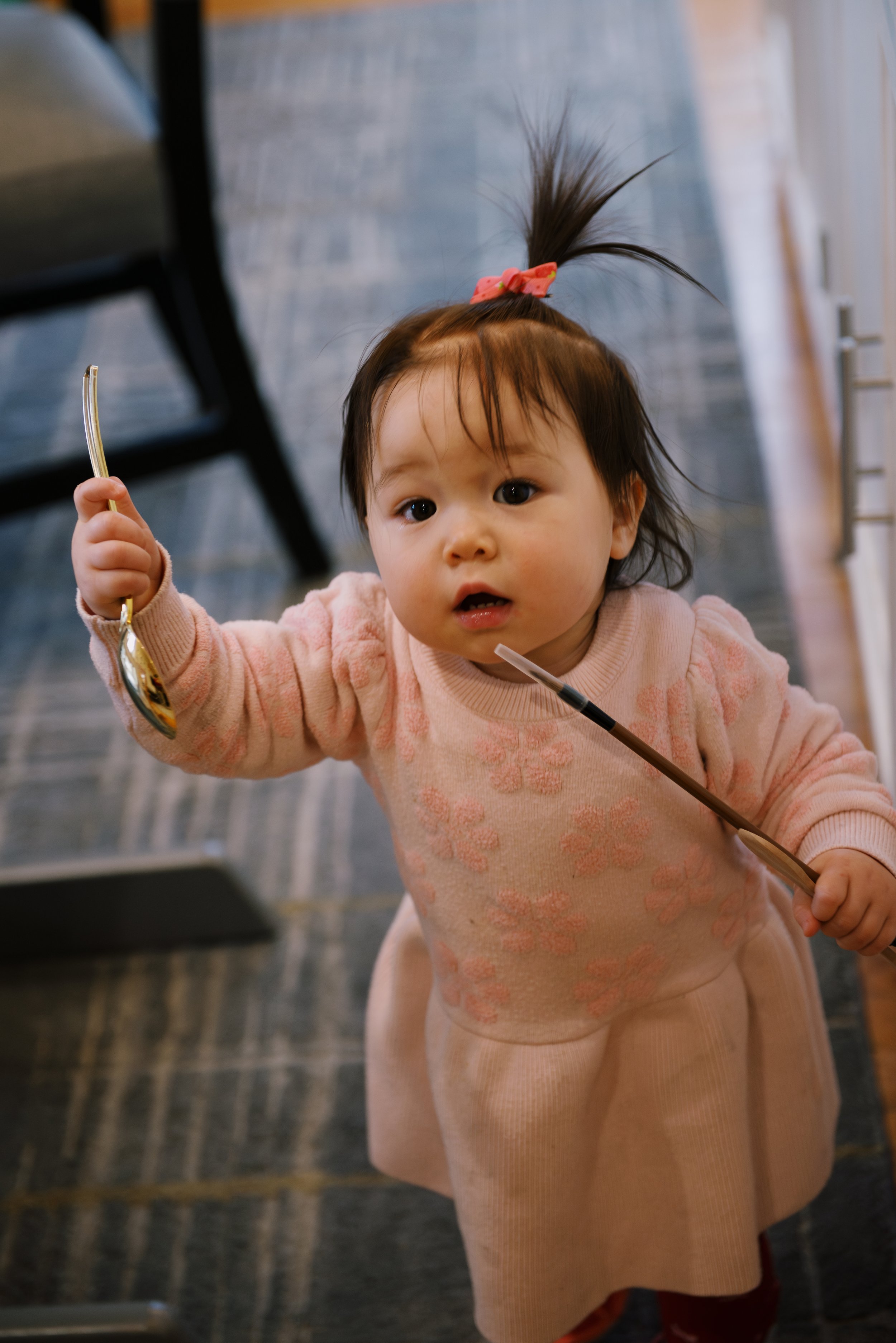 A young Asian girl with a pink bow in her hair, holding a spoon in her right hand and a pair of chopsticks in her left, looking up at the camera in a restaurant or dining area.