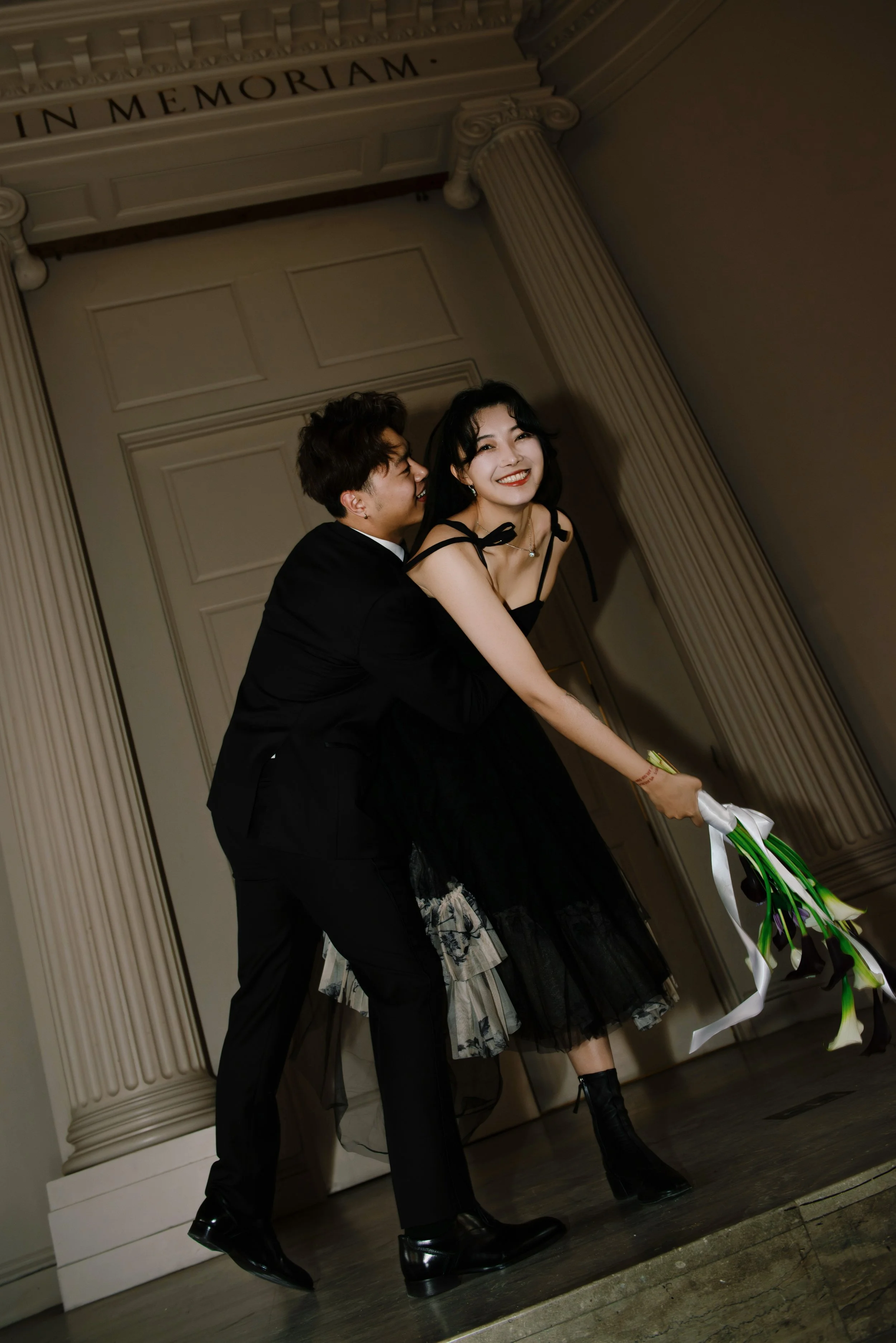 A couple dressed in black, with the woman holding a bouquet of calla lilies, dancing and smiling at an indoor event with classical columns and an archway.