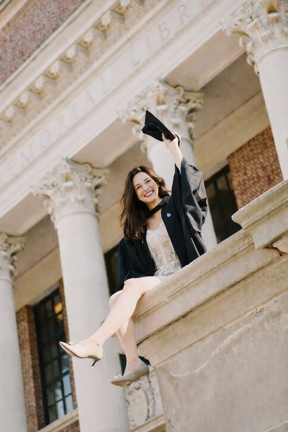 Young woman in graduation cap and gown holding a bouquet of flowers outside a building of Harvard campus with glass doors and the word 'LARAMON' on the glass.

