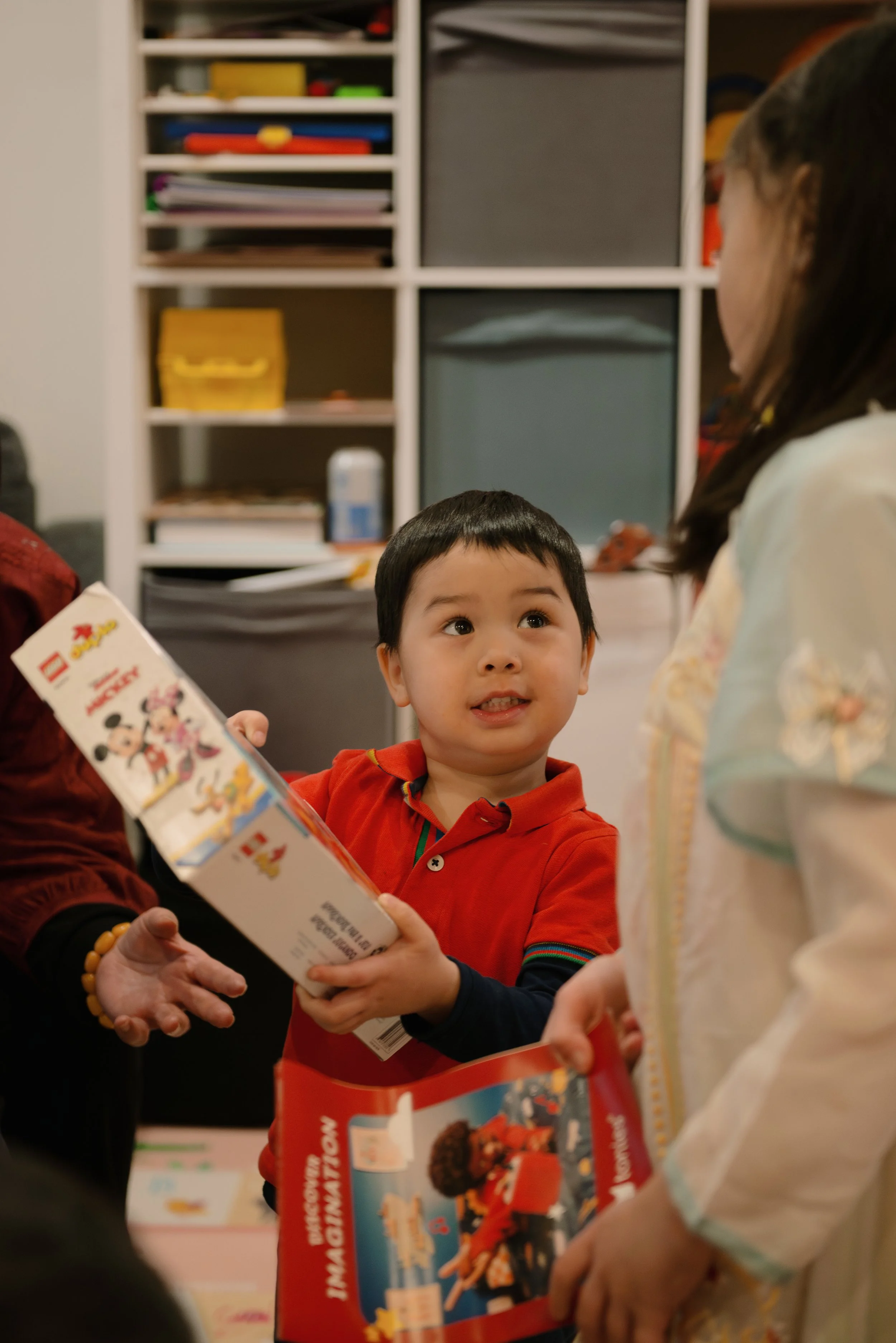 A young boy in a red shirt exchanging LEGO sets with two women at a table, in a room with a white shelving unit in the background.