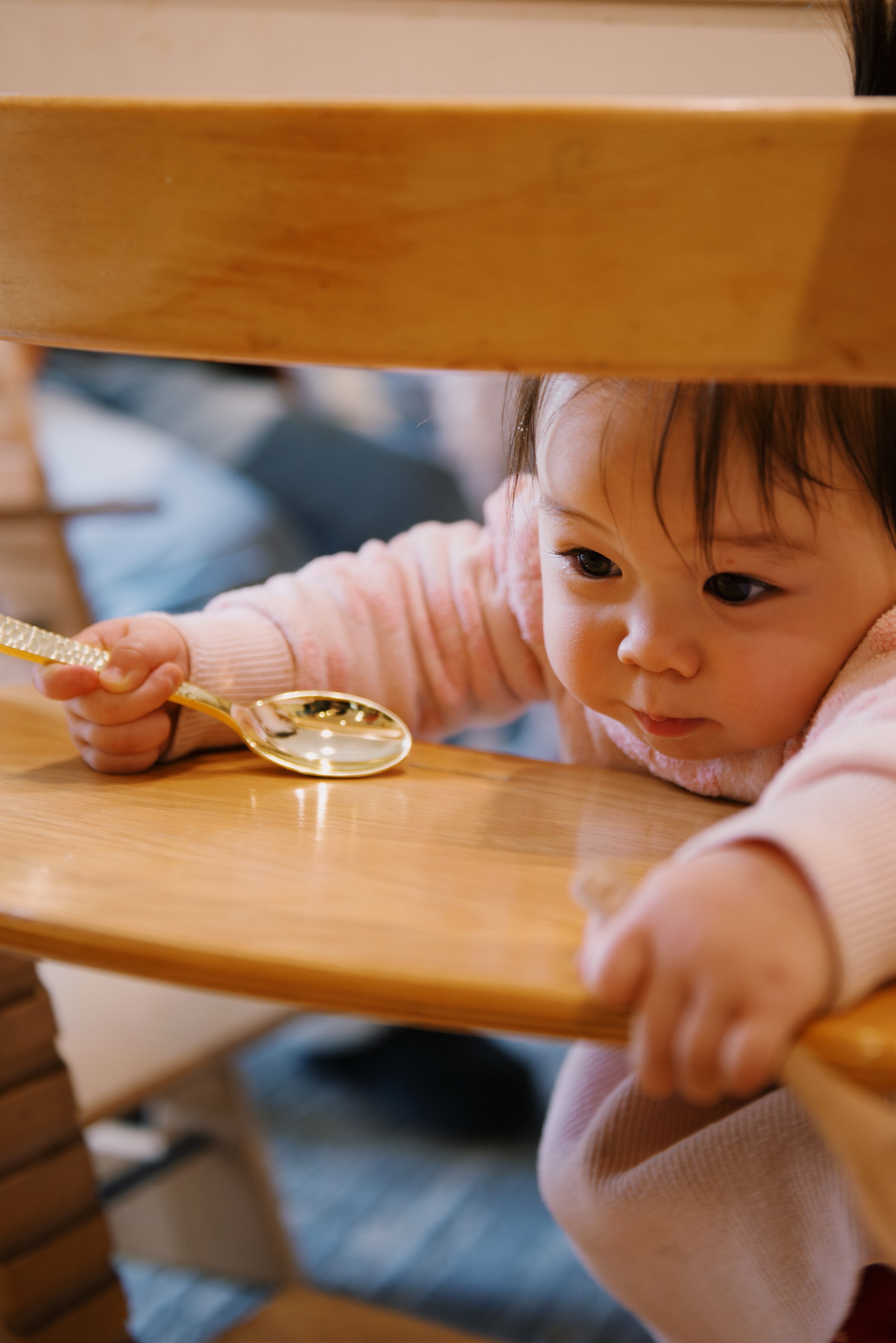 A young girl in a pink sweater looks underneath a wooden table while holding a gold spoon.