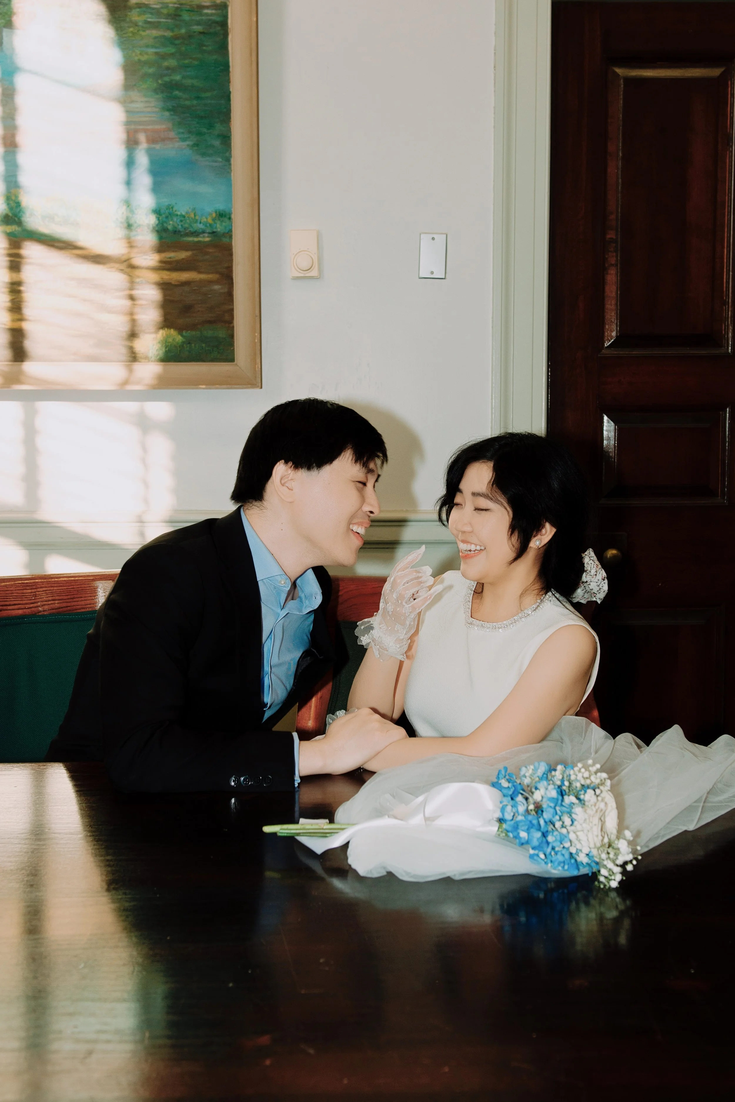 A man and woman sit at a dark wooden table, smiling and holding hands in a warmly lit room. The woman wears a white dress and lace gloves, with a bouquet of blue and white flowers on the table. The man wears a black suit and blue shirt. They are enjo