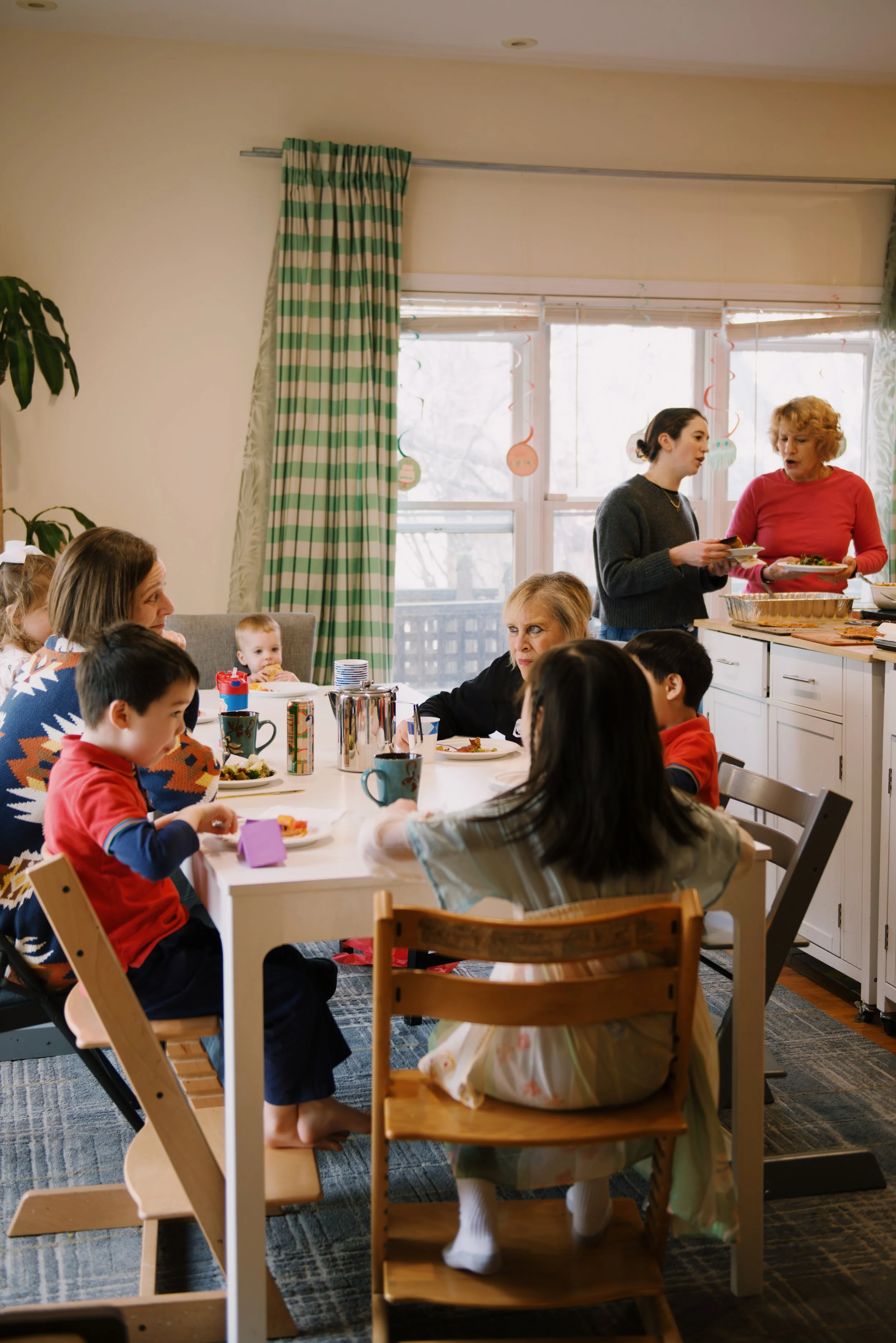 Children and adults gather around a dining table, with two women serving food in a bright kitchen with large windows and green striped curtains.