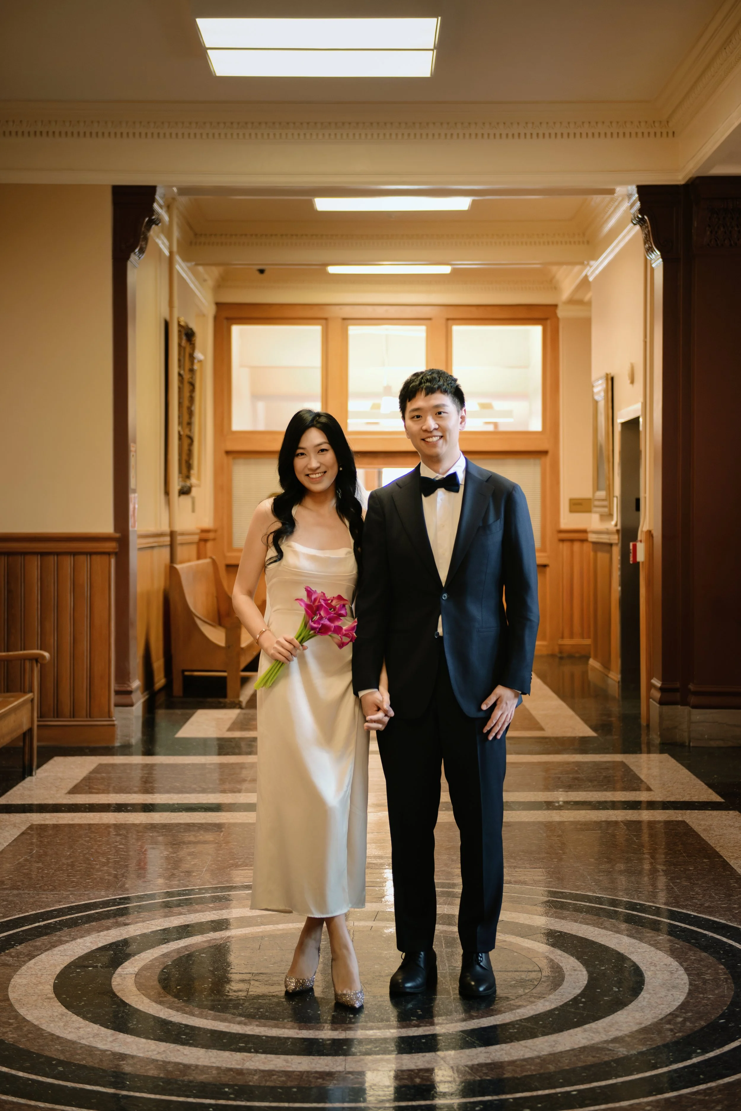 A couple dressed in formal attire posing indoors. The woman is wearing a white dress and holding pink flowers, while the man is wearing a black tuxedo with a bow tie.