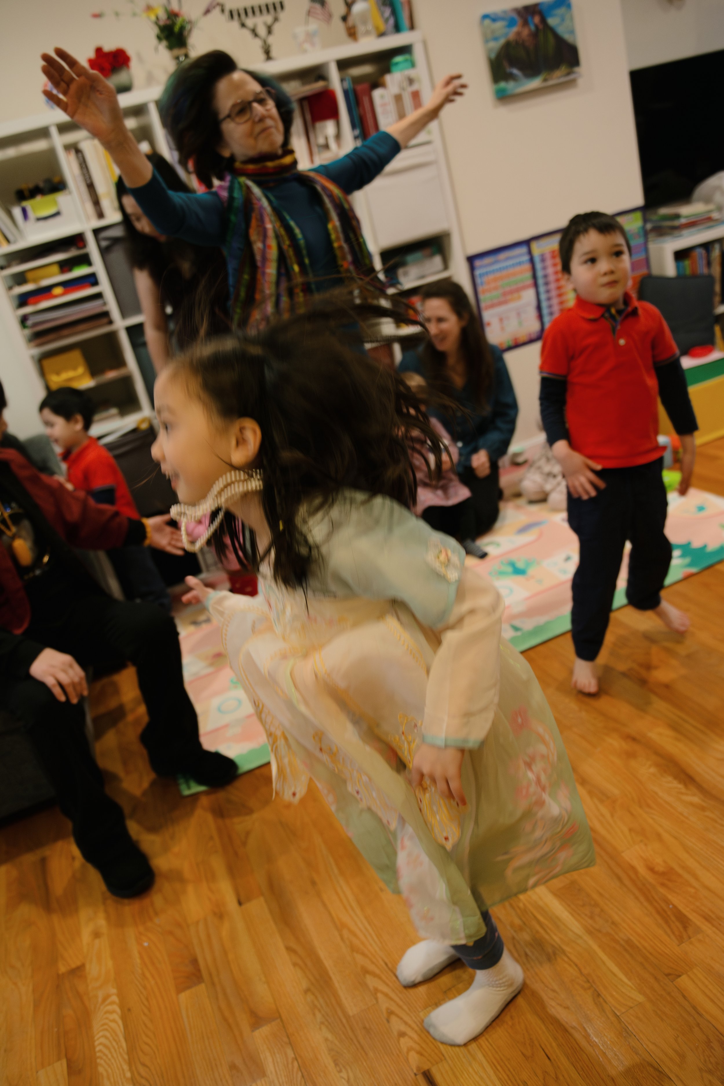 Children and adults dancing and playing in a living room with bookshelves and artwork in the background.