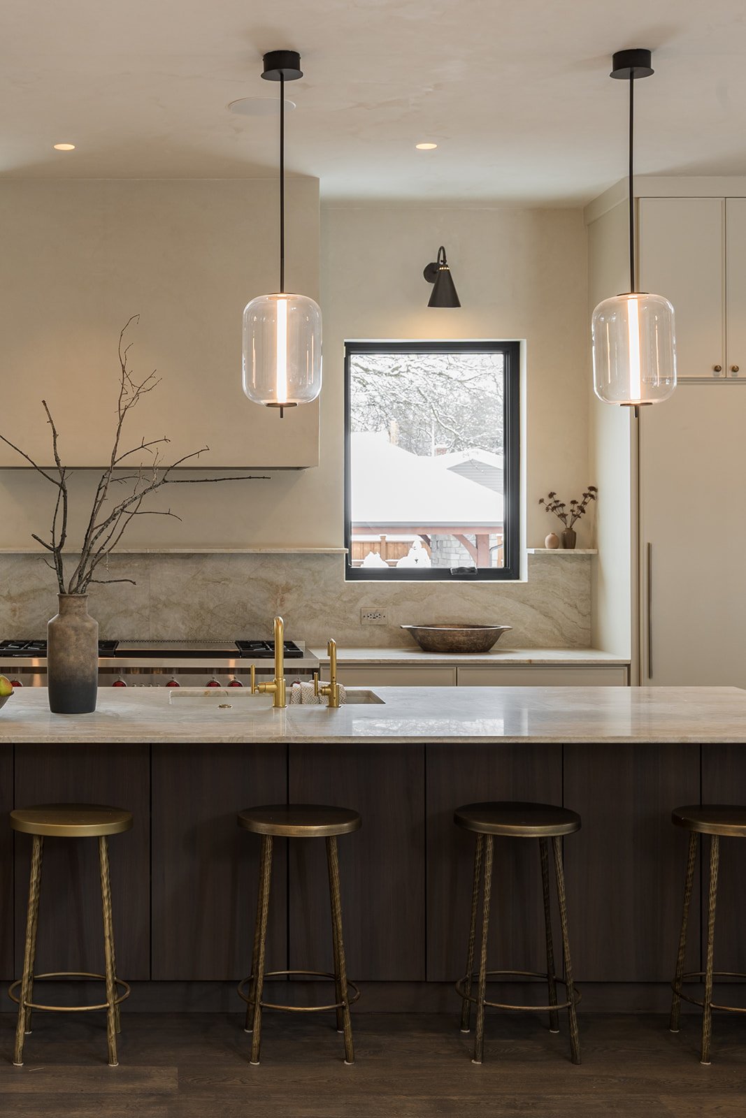Modern kitchen with white cabinetry, marble countertops, two pendant lights, and a large window showing snow outside. There are three stools at the island, a vase with branches, and some decorative items.