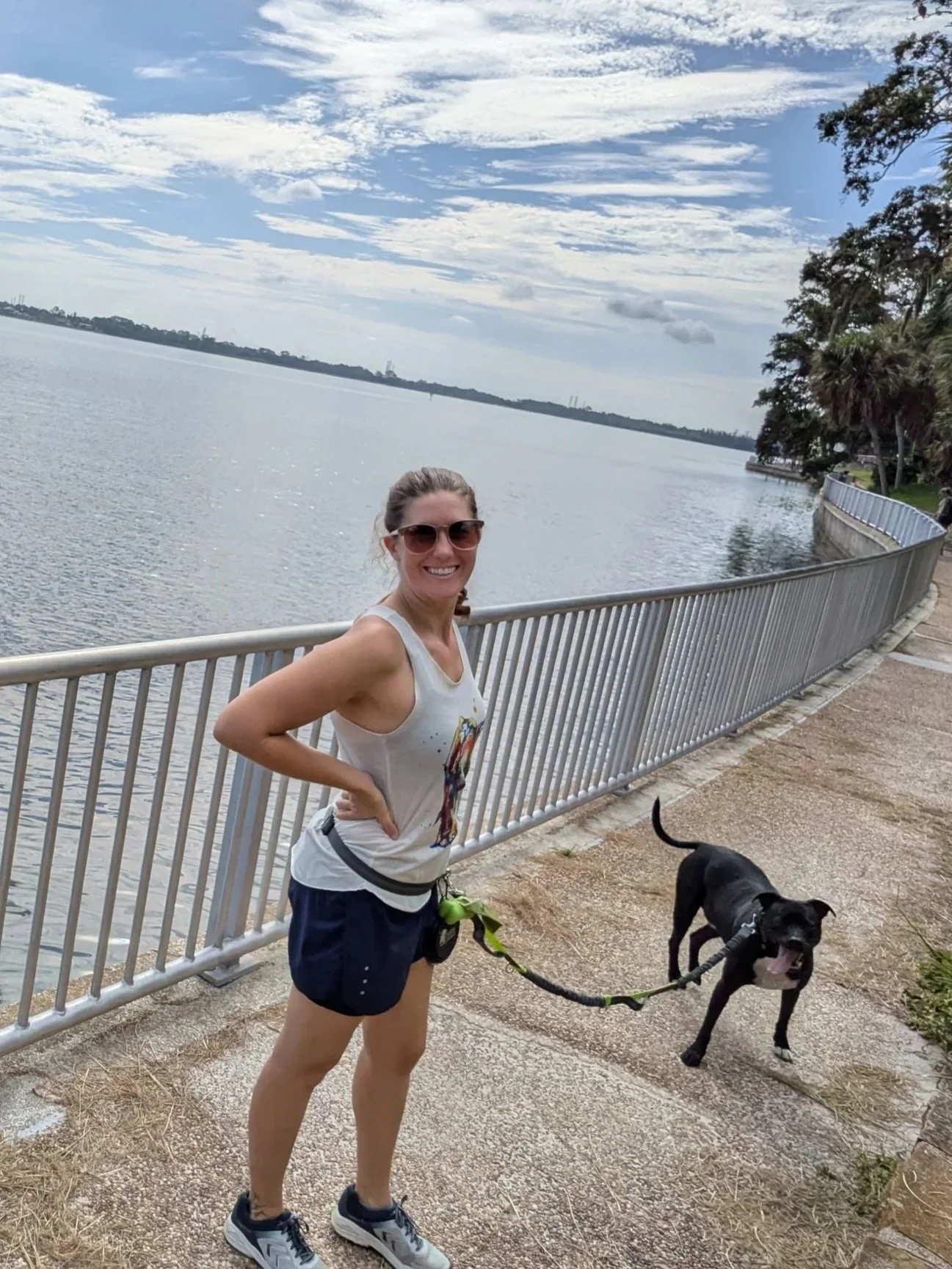 A woman wearing sunglasses and athletic clothing stands next to a black dog on a leash, smiling at the camera by a body of water with a railing and trees in the background.