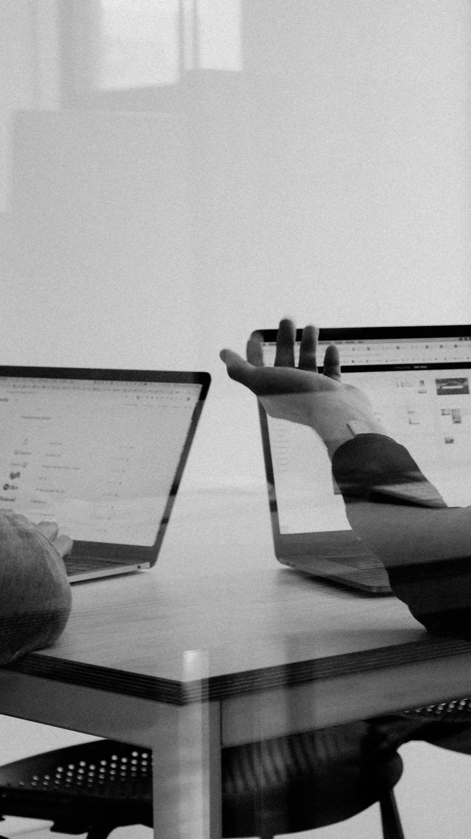 Two laptops on a desk with a person’s hand reaching towards one of the screens, in a grayscale photo.