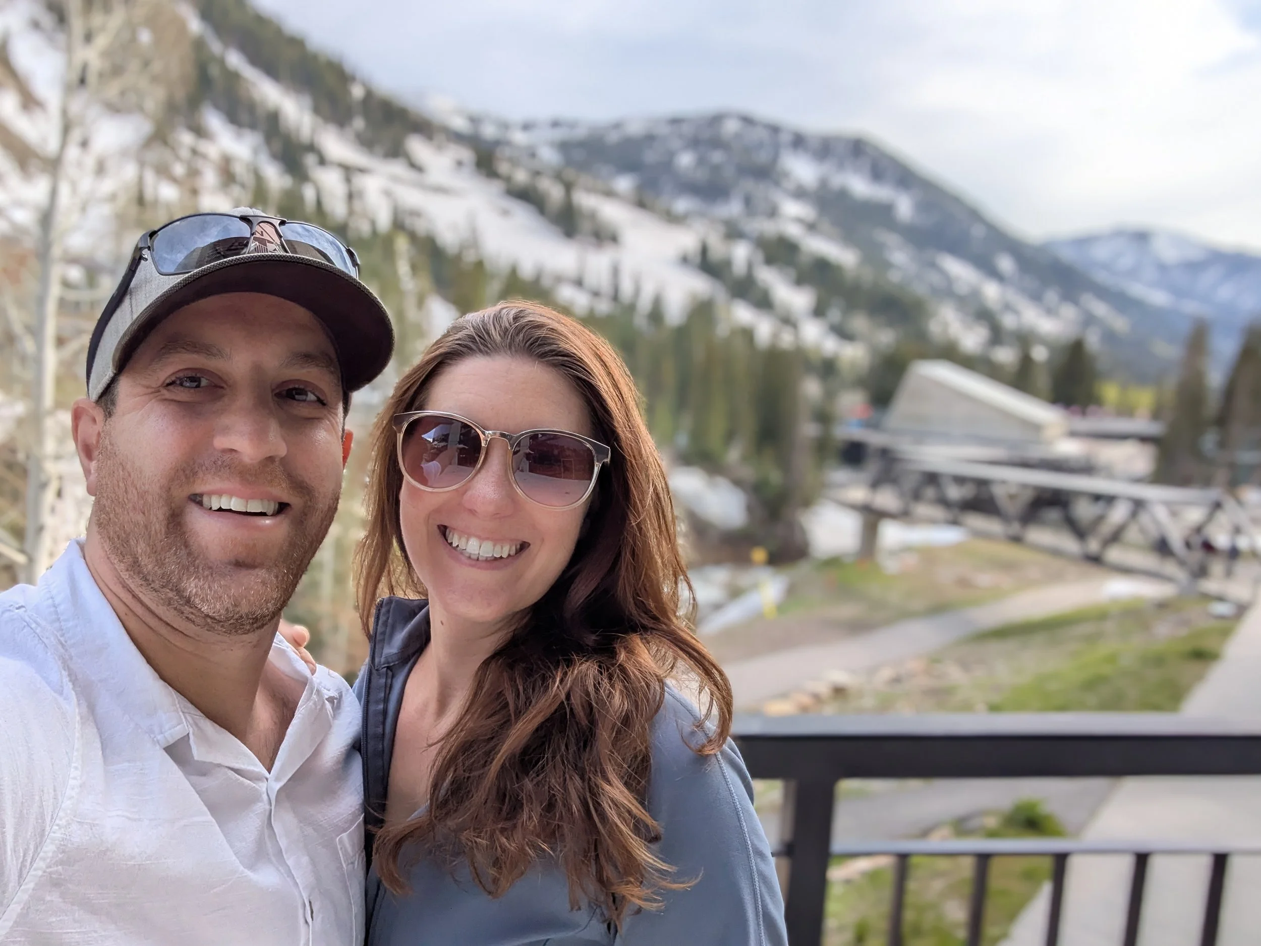 A smiling man and woman taking a selfie outdoors with snow-covered mountains in the background, woman wearing sunglasses and a jacket, man wearing a cap and sunglasses on his head.