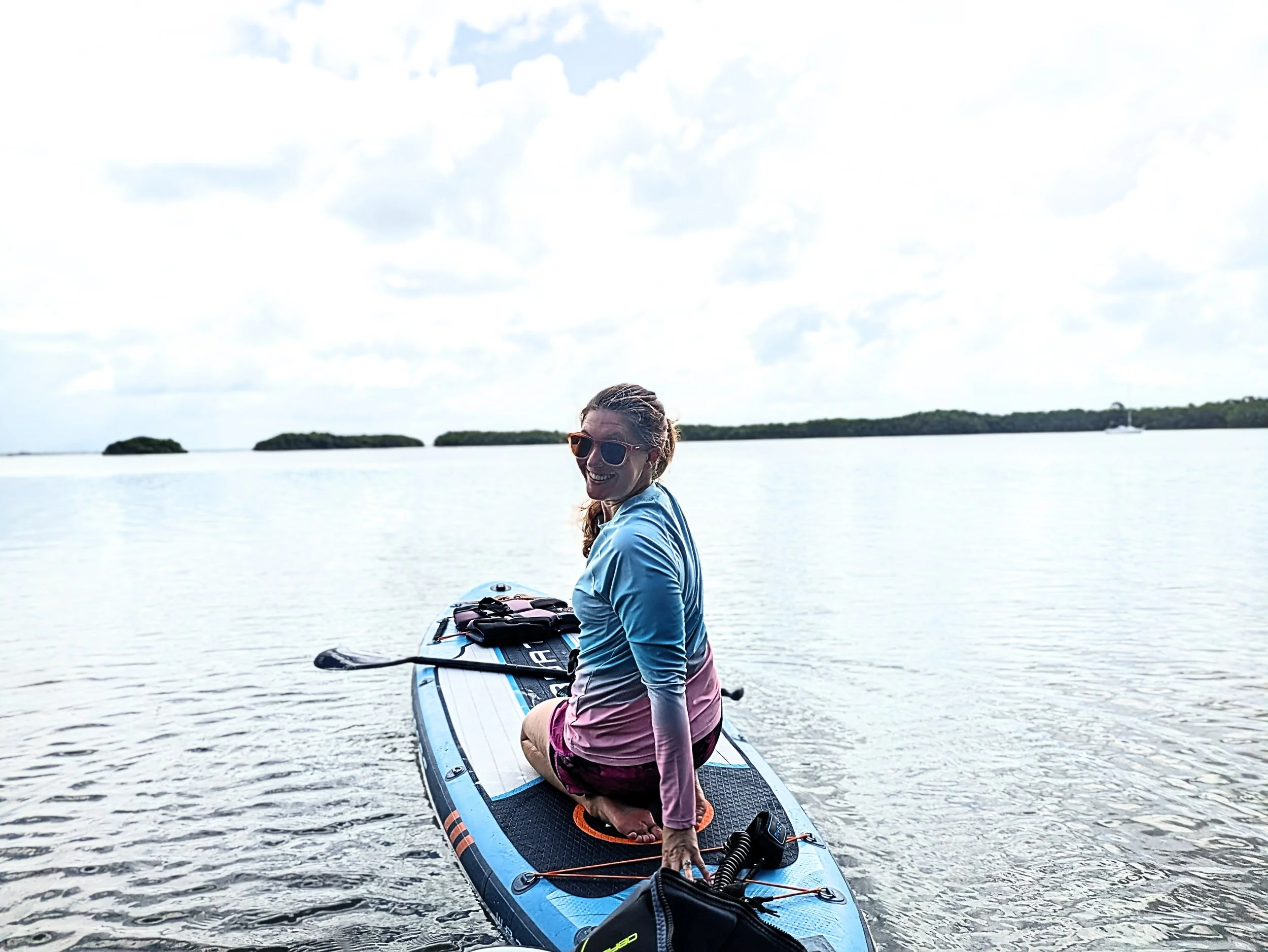 A woman sitting on a paddleboard in a calm body of water, wearing sunglasses and a long-sleeved shirt, with islands and a sailboat in the background on a cloudy day.