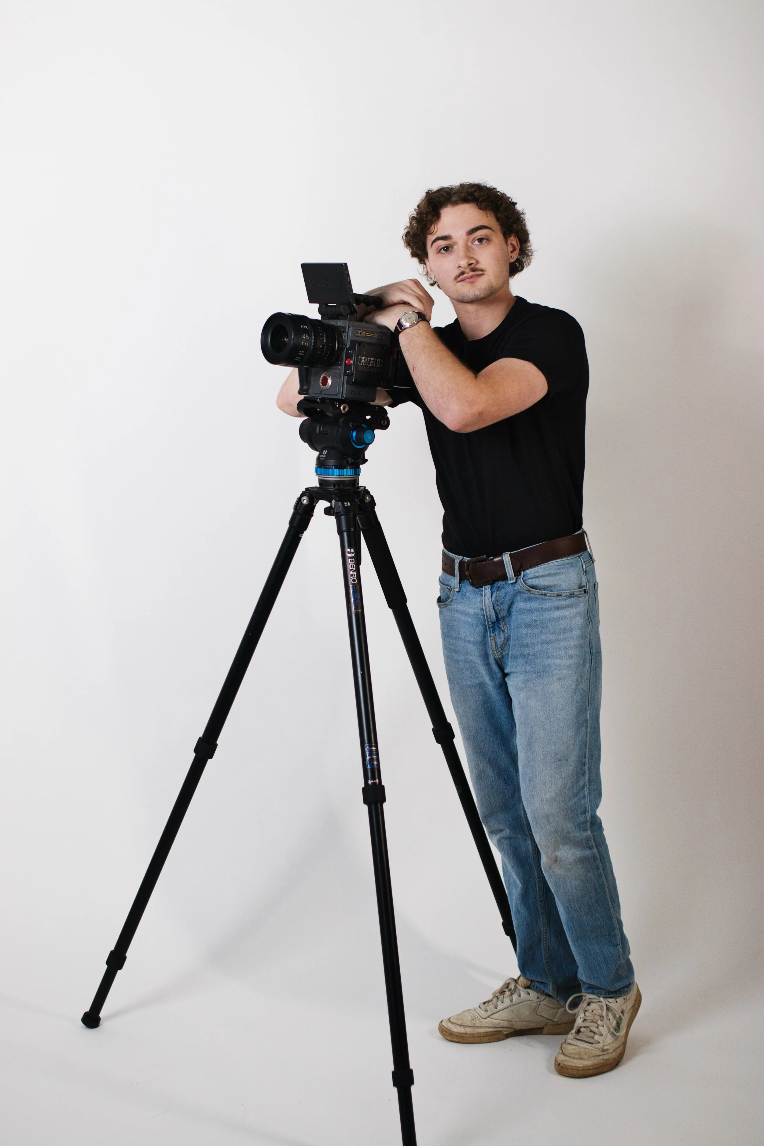 A young man with curly hair and a mustache standing next to a professional video camera on a tripod, looking at the camera with a neutral expression, wearing a black t-shirt, light blue jeans, and white sneakers, against a plain white background.