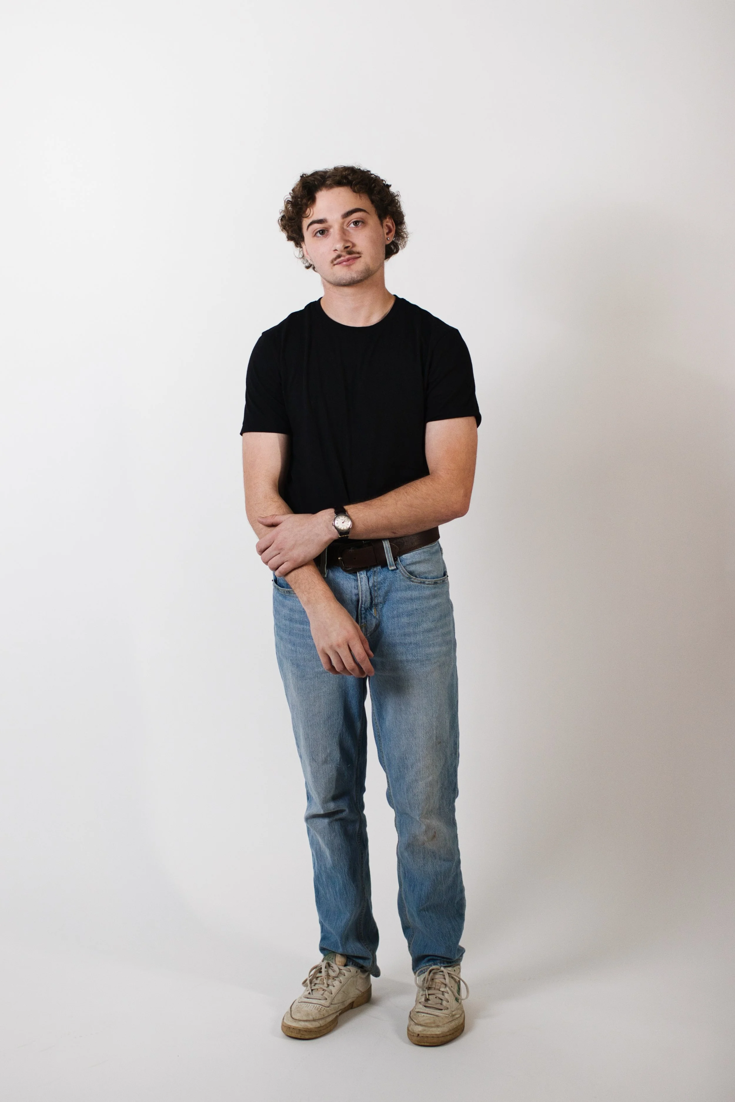 A young man with curly hair wearing a black t-shirt, blue jeans, and beige sneakers standing against a white background.