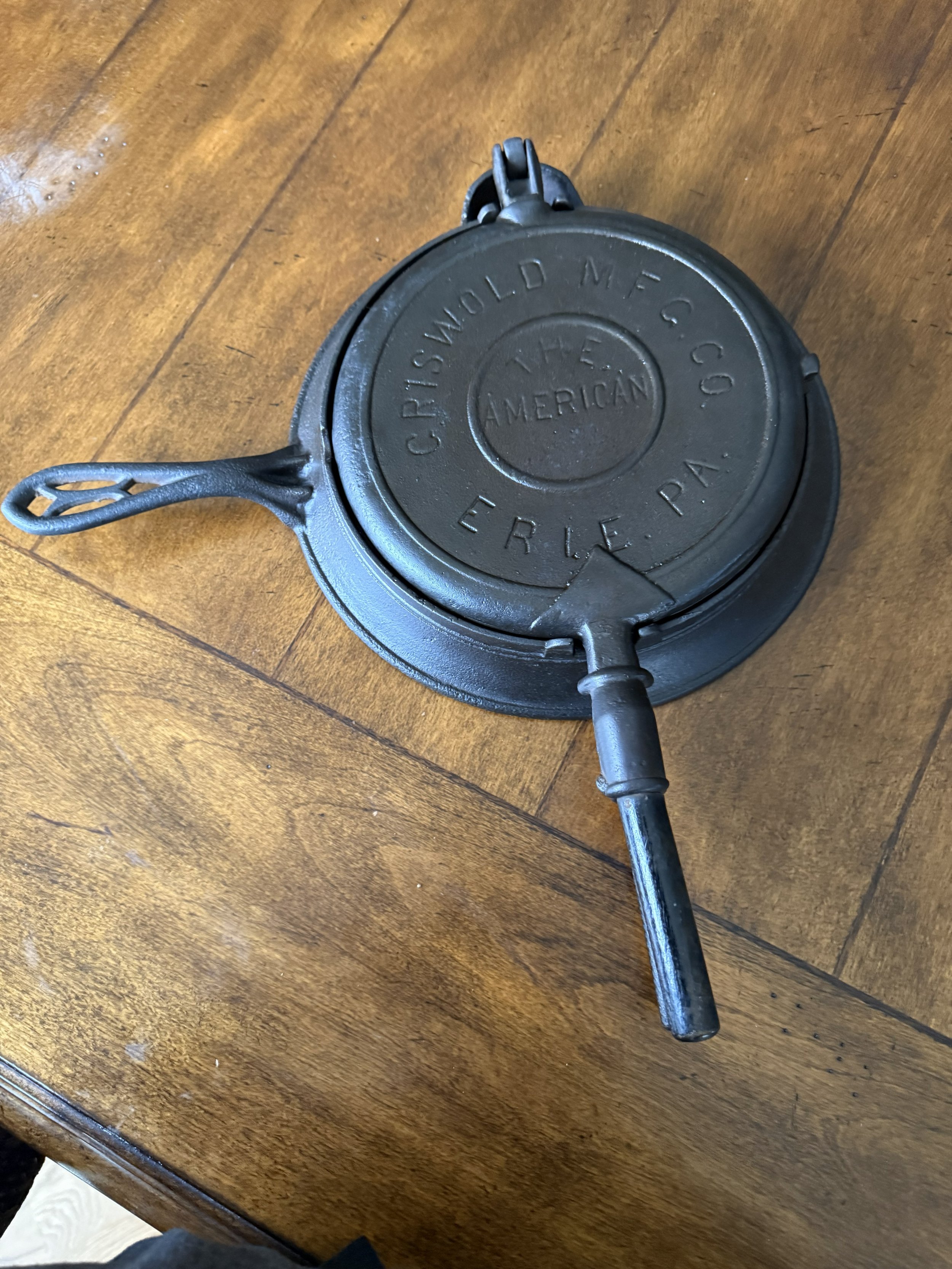 Vintage black cast iron corn bread mold with the words 'Crisp T. The American' and 'Erie PA' engraved on the lid, resting on a wooden surface.