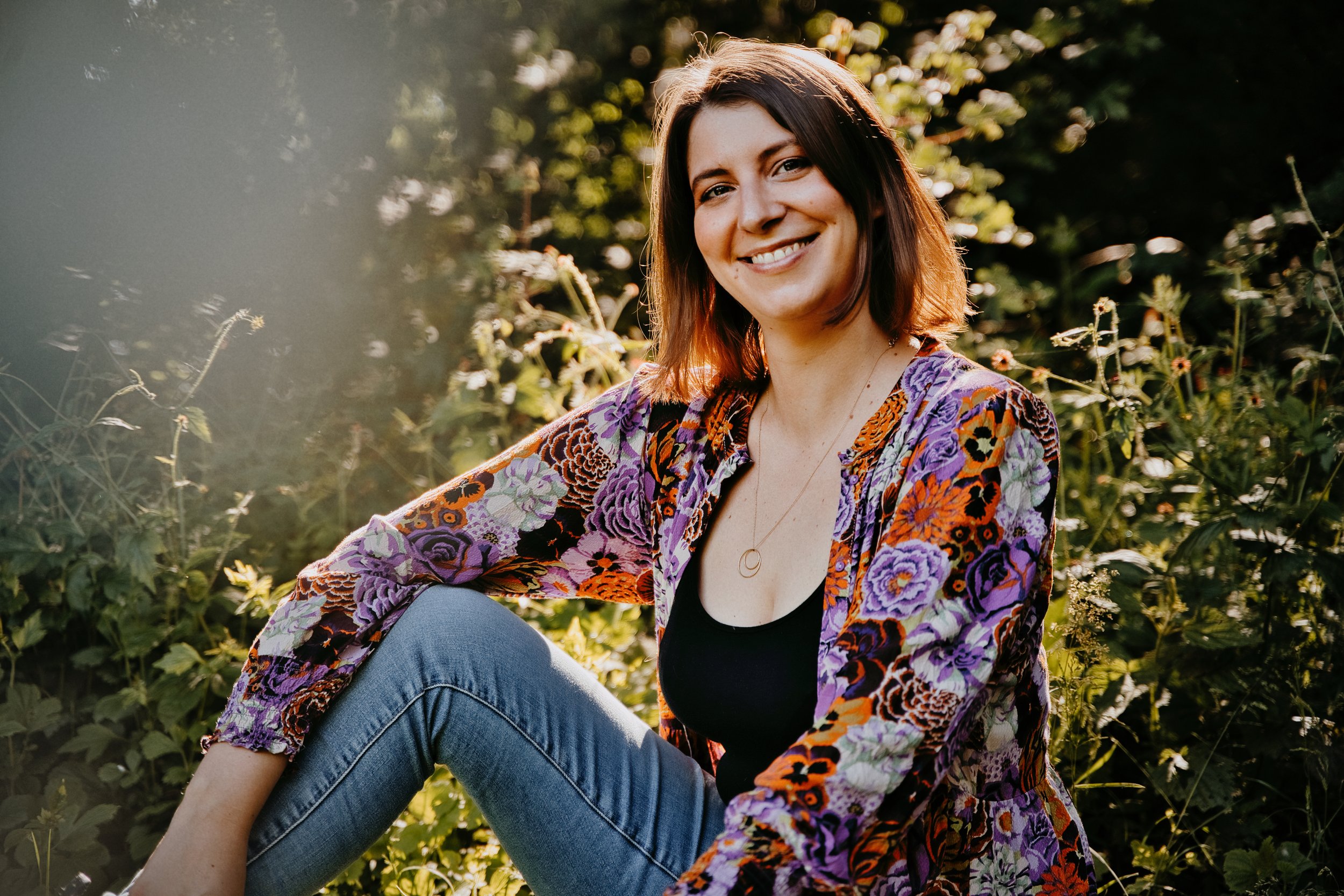 Lisa, a femme human, smiling outdoors surrounded by greenery, wearing a floral jacket and blue jeans, sitting on the ground with sunlight in the background.