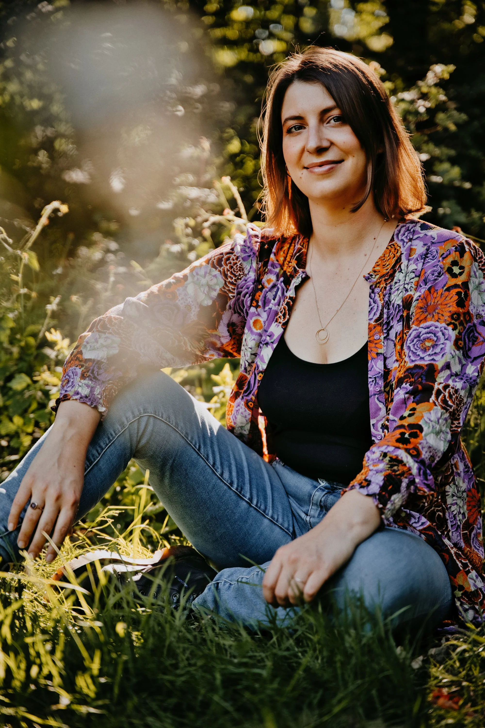 Lisa, a femme human, sitting outdoors in a grassy area during golden hour, wearing a floral patterned jacket, black top, and jeans.