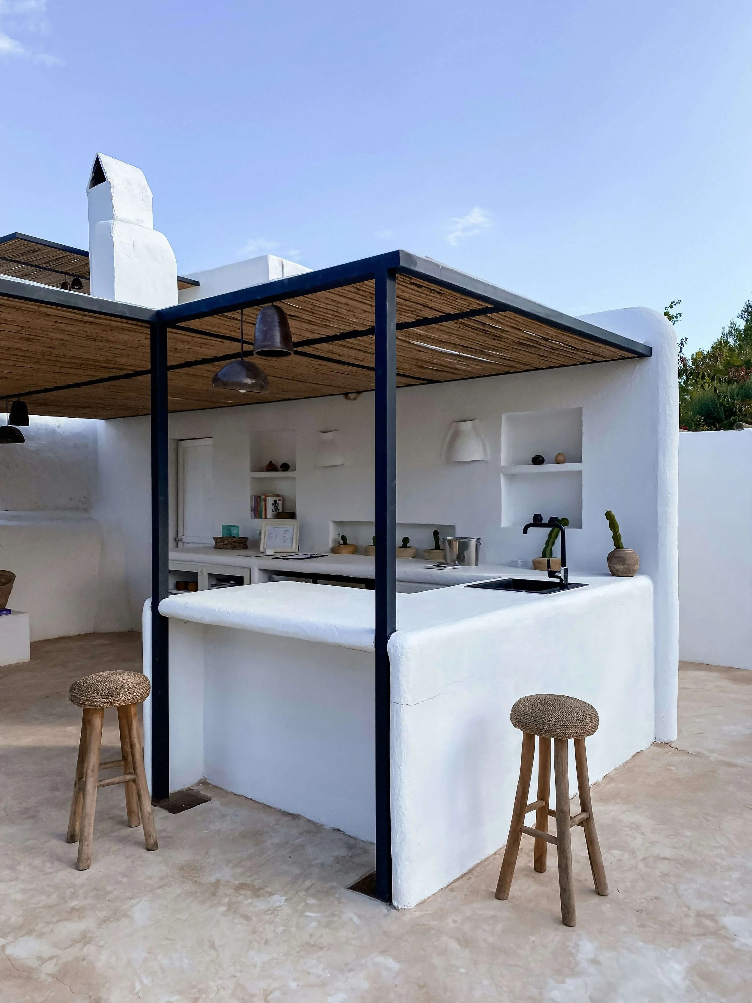 White outdoor kitchen with black frame, wooden ceiling, and two wooden stools on a concrete floor, set against a white stucco wall under a clear blue sky.