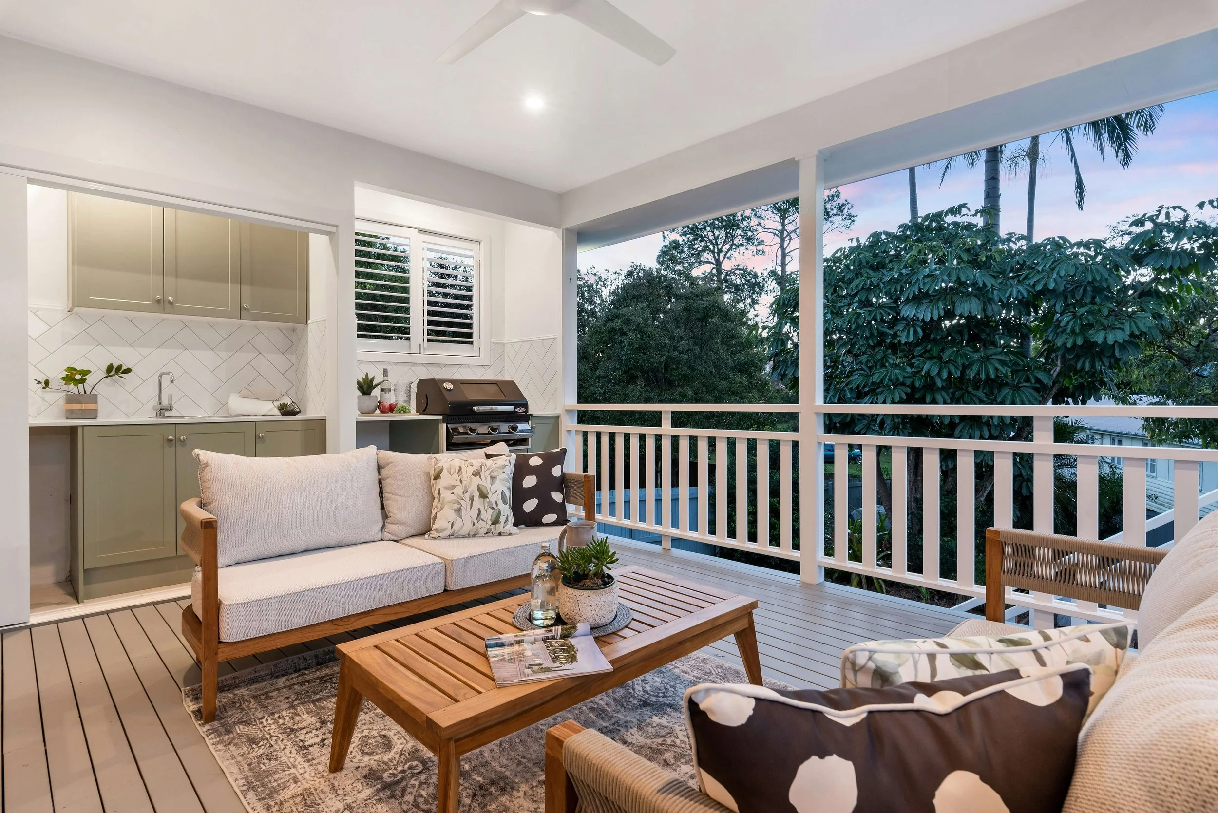 Covered balcony with white railing, cozy outdoor seating area with beige and patterned cushions, wooden coffee table with decorative items and magazine, small outdoor kitchen with gray cabinetry, grill, plants, and window with shutters, overlooking trees at dusk.
