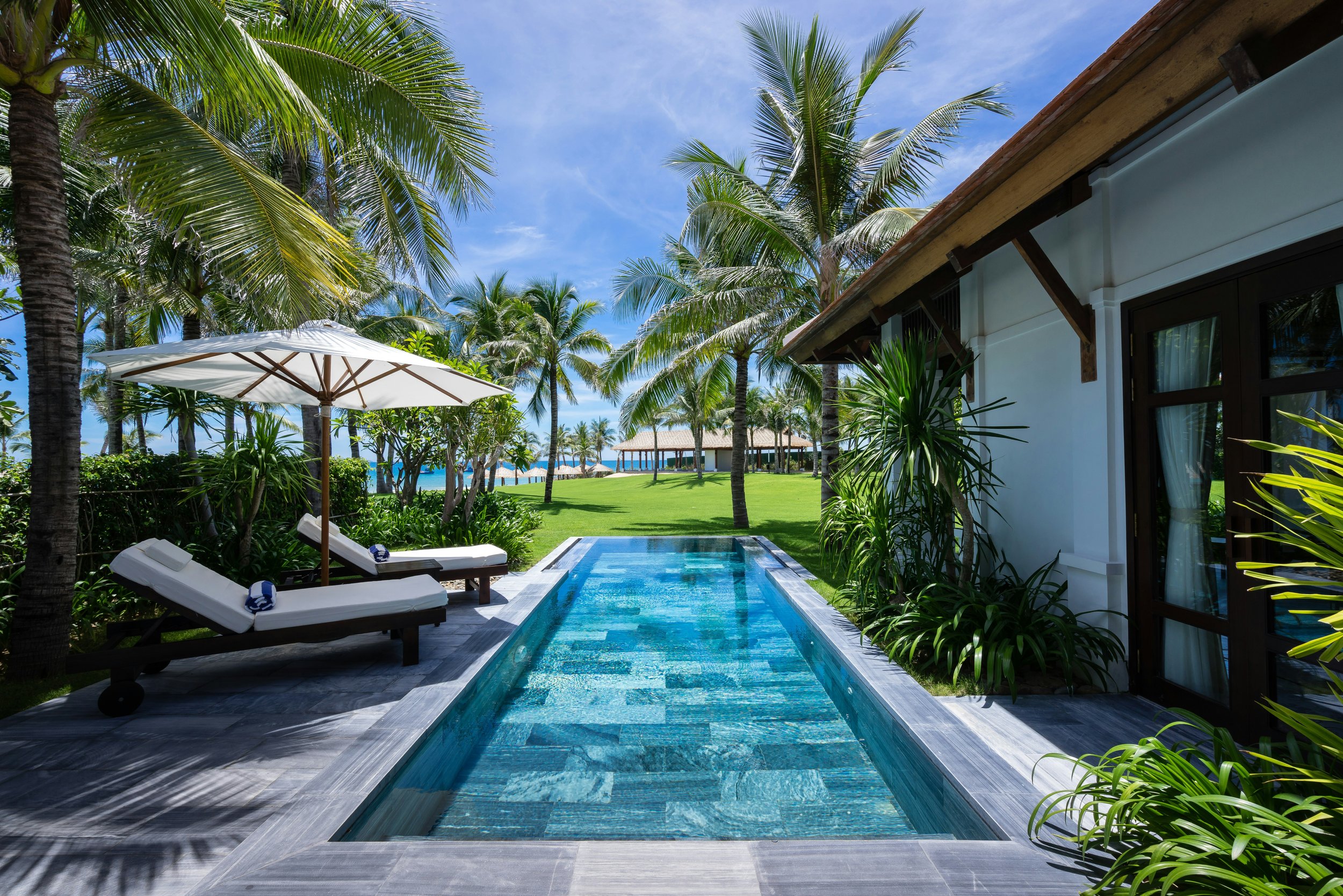 A luxurious poolside scene at a tropical resort with lounge chairs, umbrella, lush greenery, tall palm trees, a well-maintained lawn, and a view of the ocean in the background under a bright blue sky.