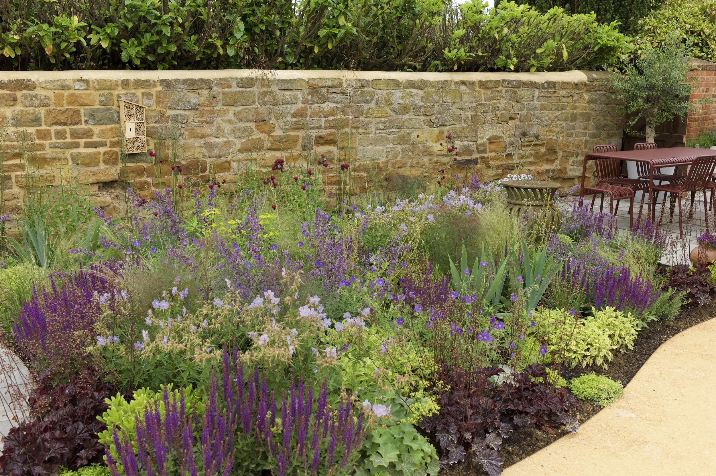Colorful garden with purple, lavender, and green plants along a stone wall and a paved patio with outdoor furniture.