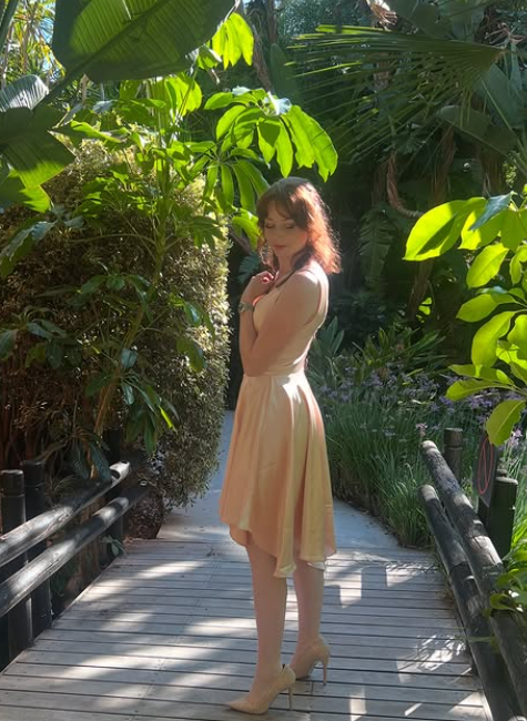 A beige ceramic vase with a narrow neck and rounded body, holding a branch with small round seed pods, sitting on a wooden surface with shadow cast on a light-colored wall.