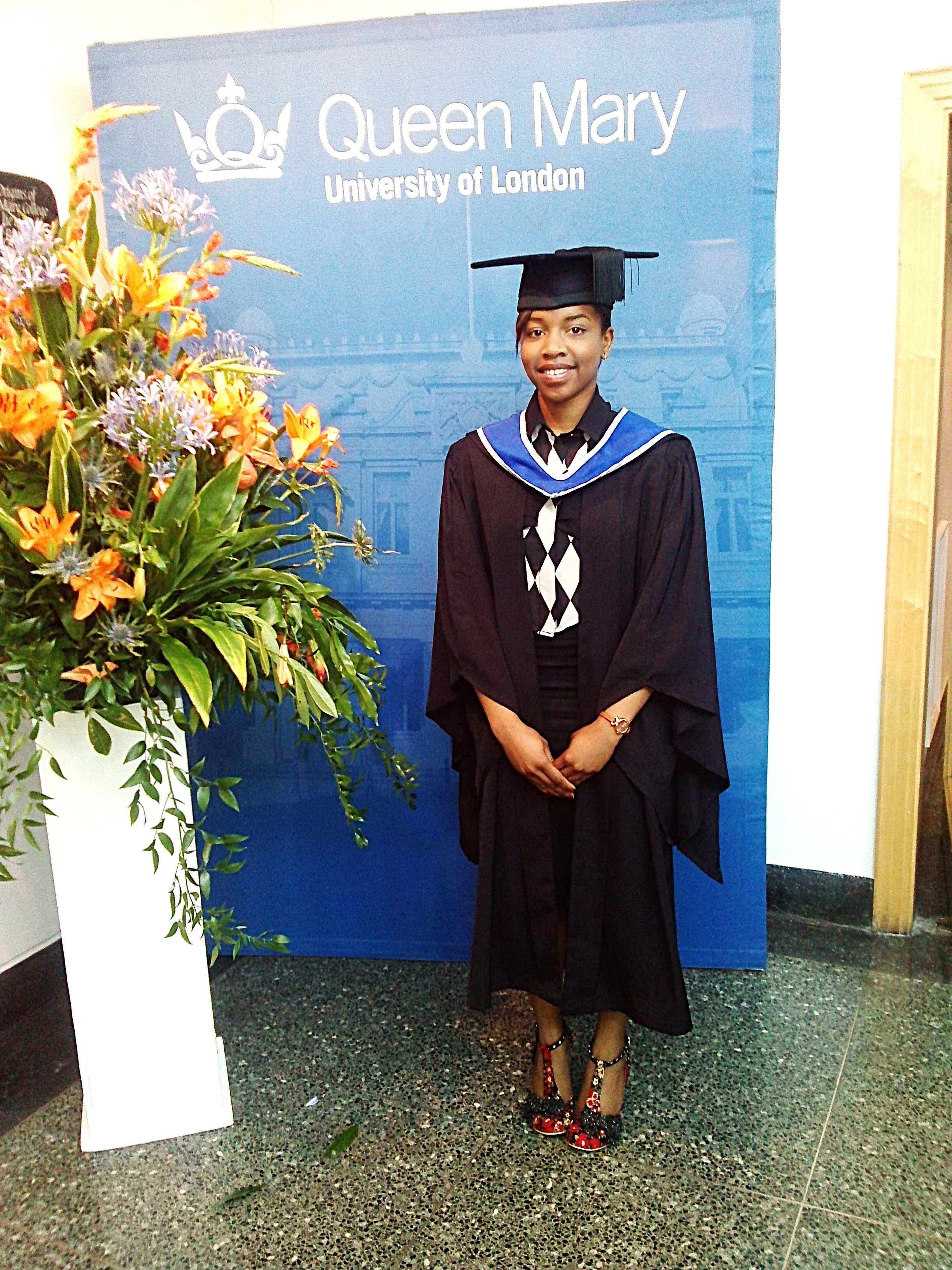Ope the Tutor in a Queen Mary University of London (Russell Group) graduation gown and cap, standing in front of a blue backdrop at the Chemistry School ceremony
