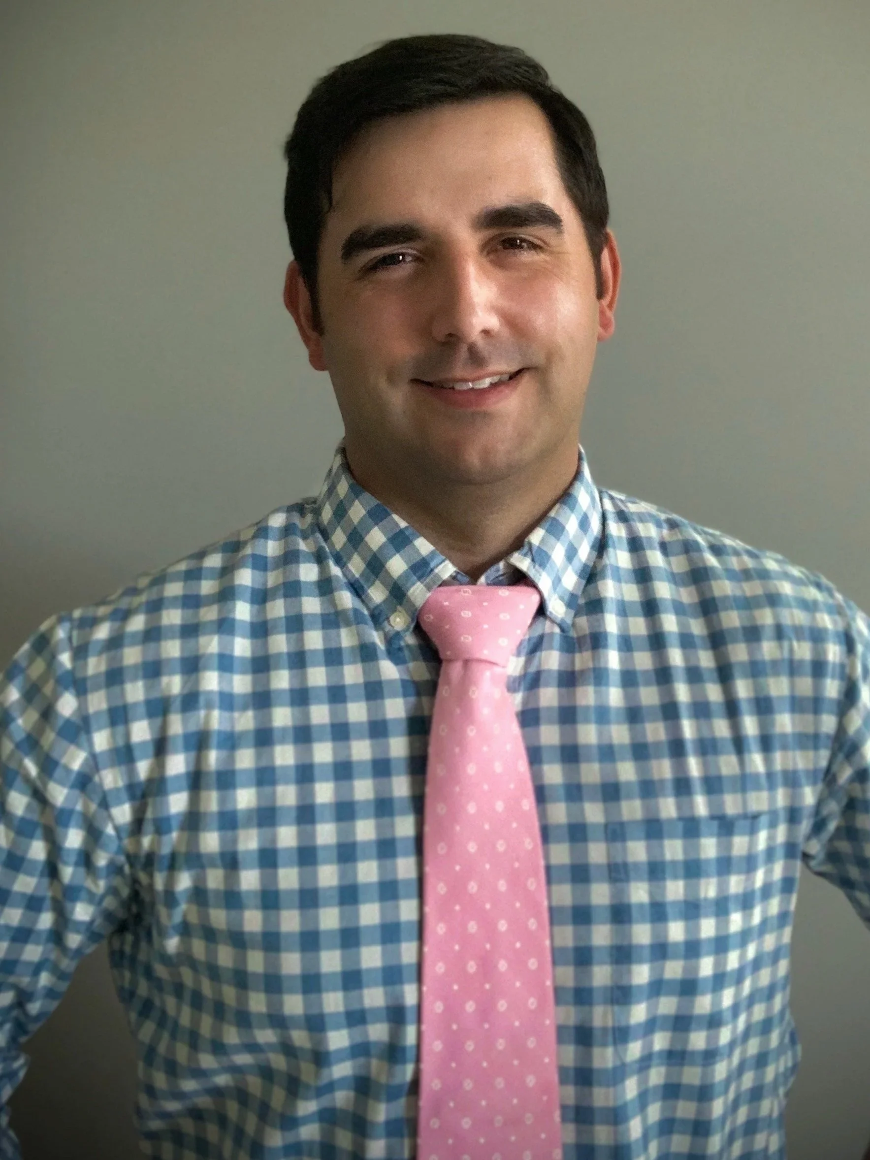 A man wearing a blue and white checkered shirt with a pink tie, standing against a plain gray background.