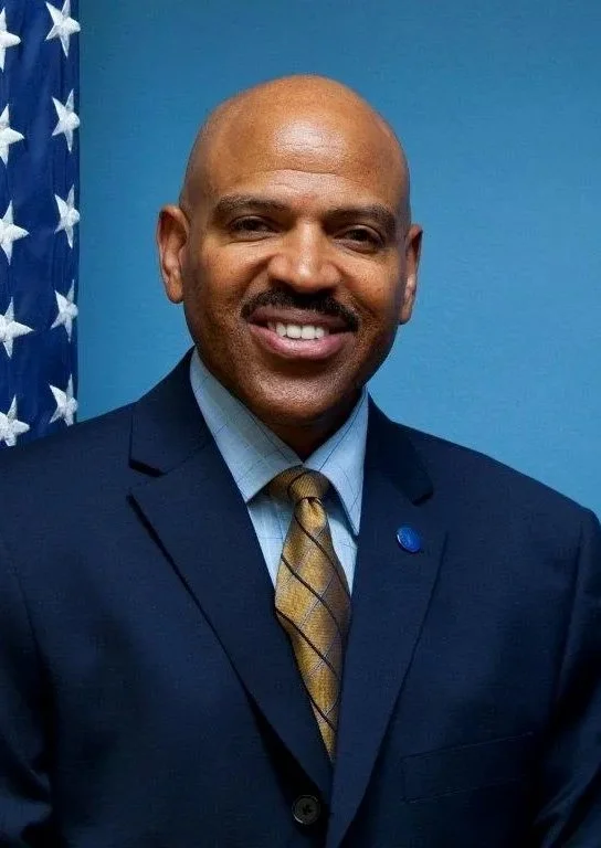 A man dressed in a navy suit, light blue shirt, and a checkered gold and blue tie, standing in front of an American flag and a blue wall, smiling at the camera.