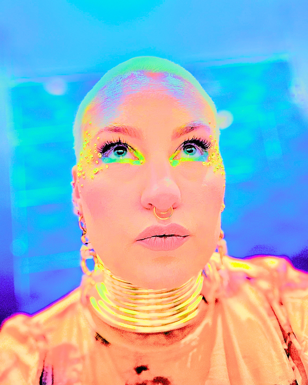 A person with rainbow-colored makeup, rhinestones on the face, multiple gold jewelry pieces, and a metallic gold top, looking upwards against a vibrant blue background.