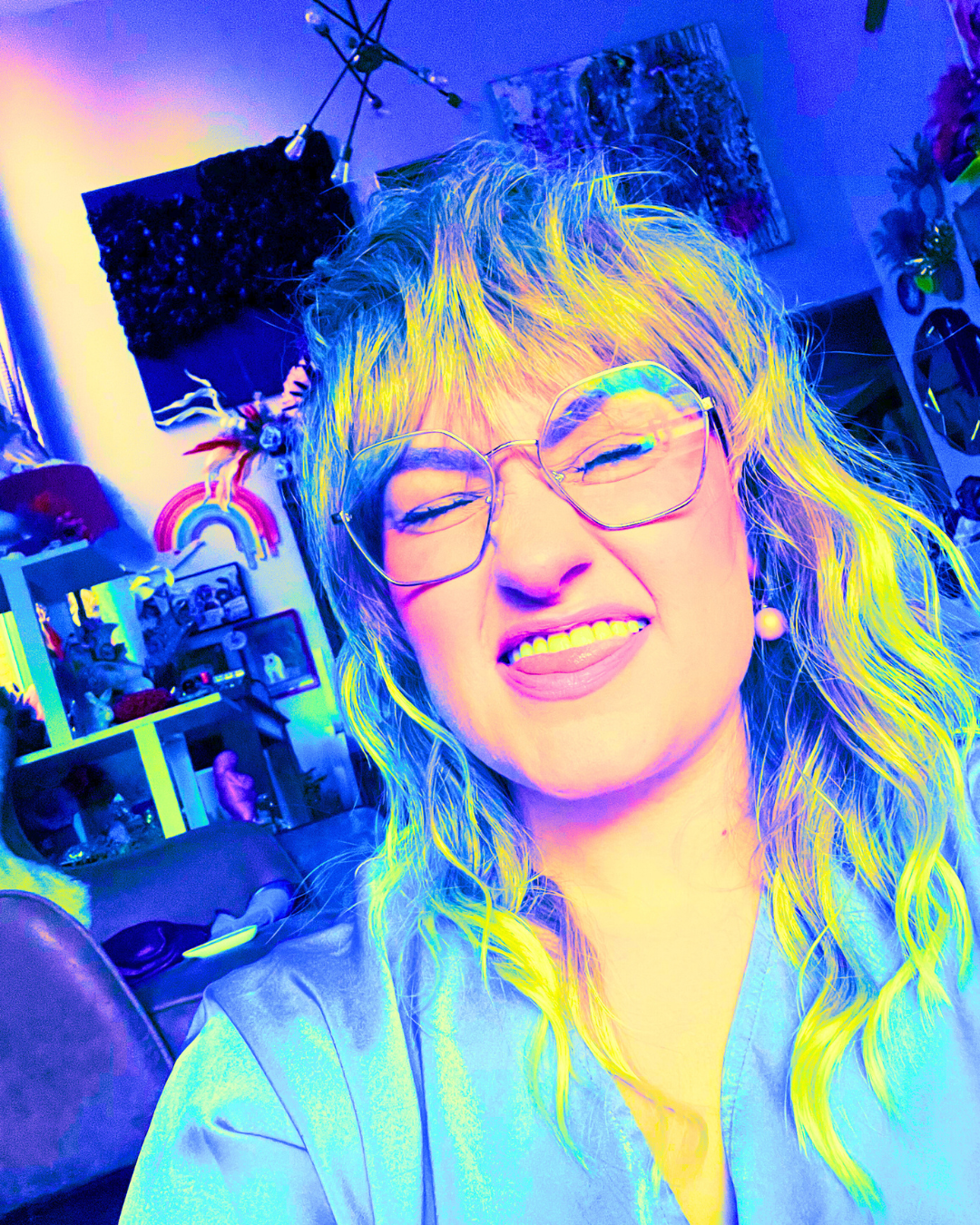 A woman with long blonde hair, wearing glasses and a light-colored top, making a playful expression with her tongue sticking out. The background shows a colorful, cluttered room with art, decorations, and furniture under vibrant blue and purple lighting.