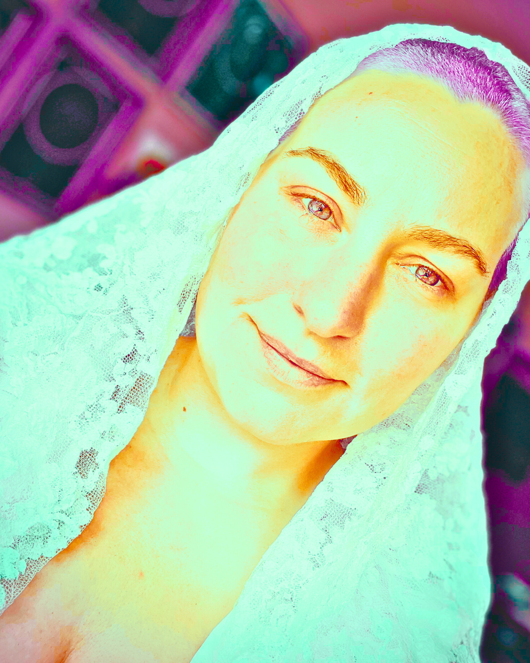 Close-up portrait of a young woman with a lace veil over her head, light skin, and light-colored eyes, smiling slightly, with a blurred background.